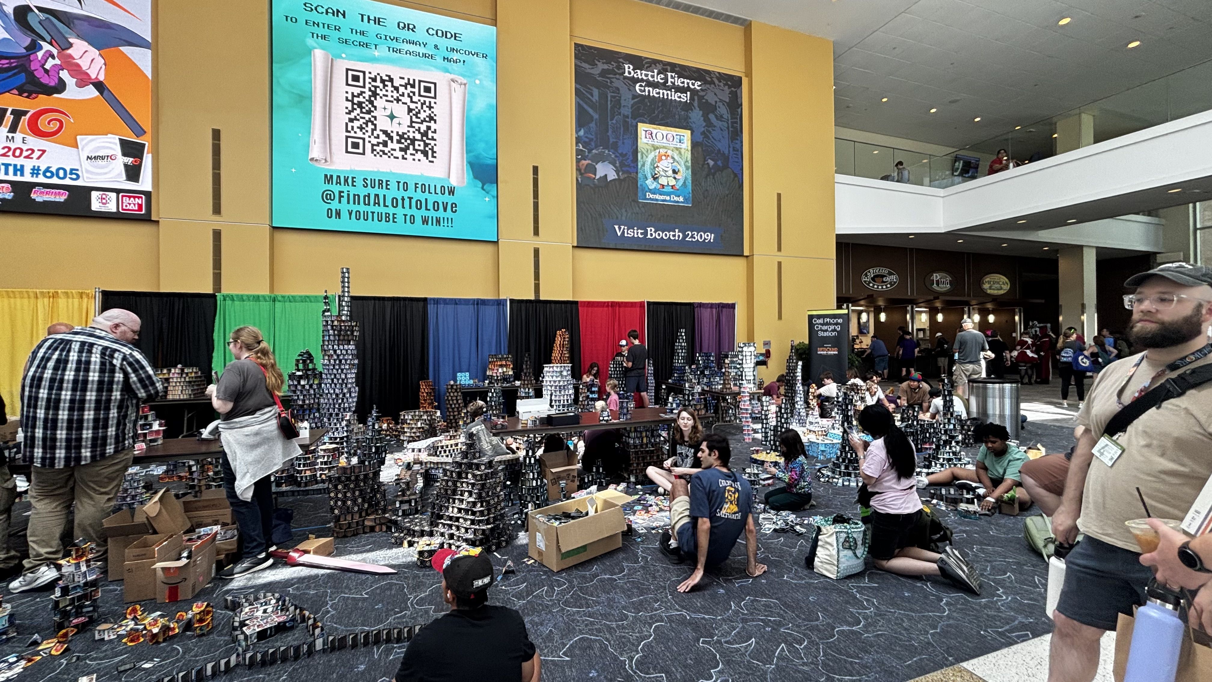 Busy convention center with yellow walls and large banners, including a teal QR-code poster. Foreground shows vendors with tall stacks of display towers while attendees browse; balcony above with people.
