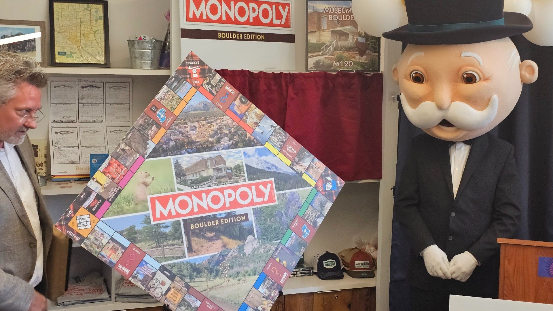 Man unveiling a large Monopoly Boulder Edition board next to a person in Monopoly Man costume with a black top hat and white gloves in a room with framed maps and memorabilia.