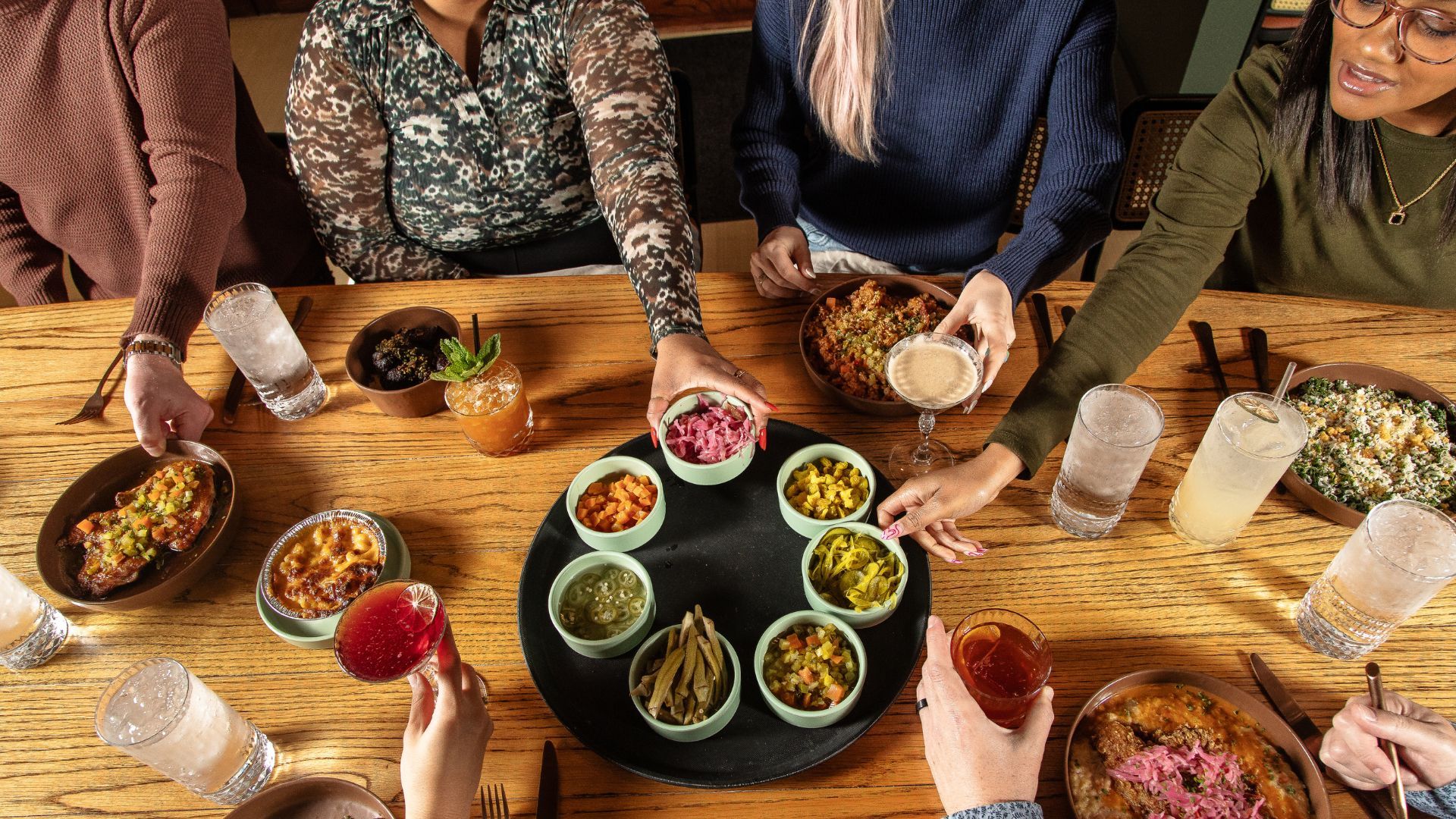 A group of people shares a meal, reaching for colorful small plates and drinks around a wooden table.