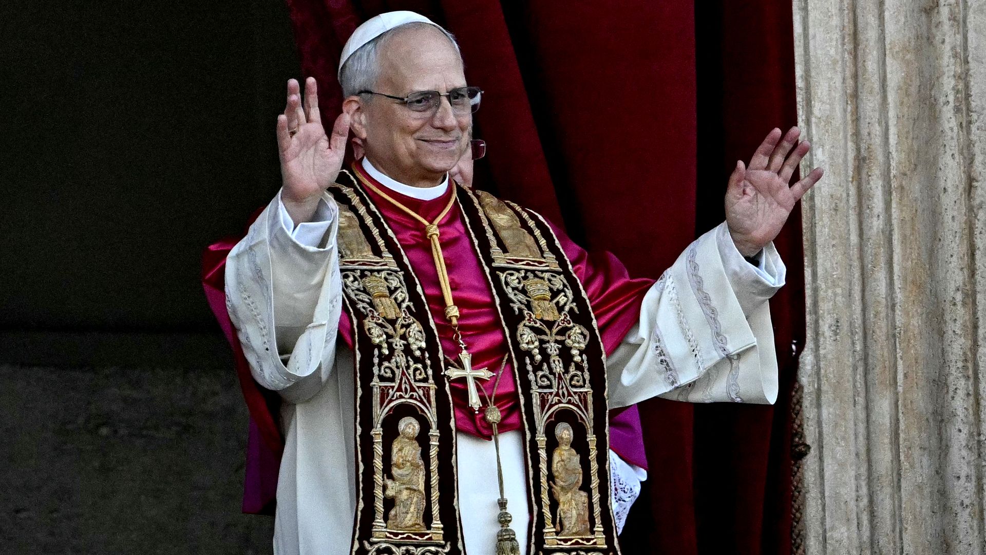 Pope Leo holds up both hands as he walks onto a balcony, smiling slightly.