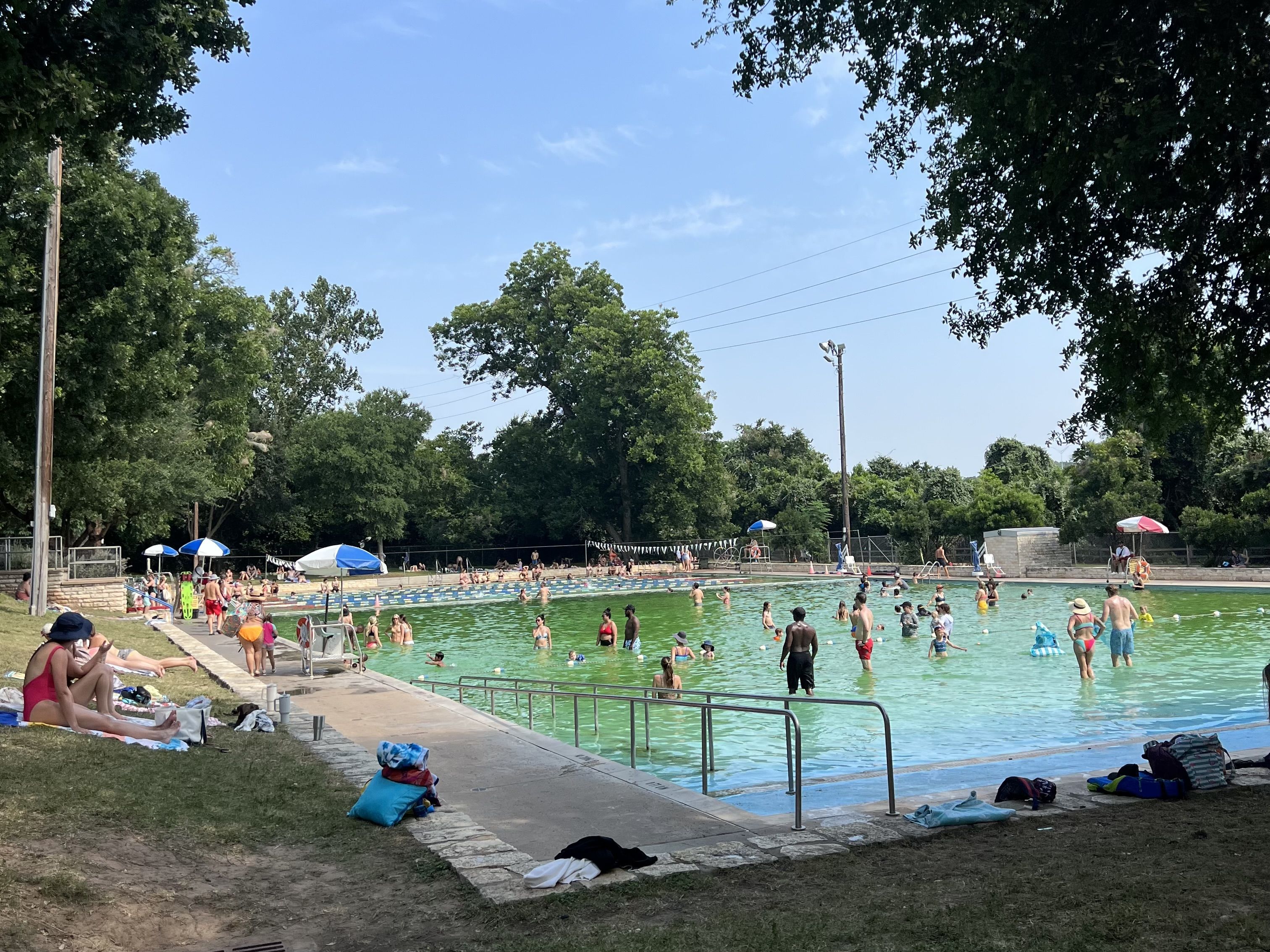 Busy outdoor swimming pool surrounded by green trees under a clear blue sky. Dozens of swimmers fill the water while sunbathers relax along the edge under blue umbrellas.