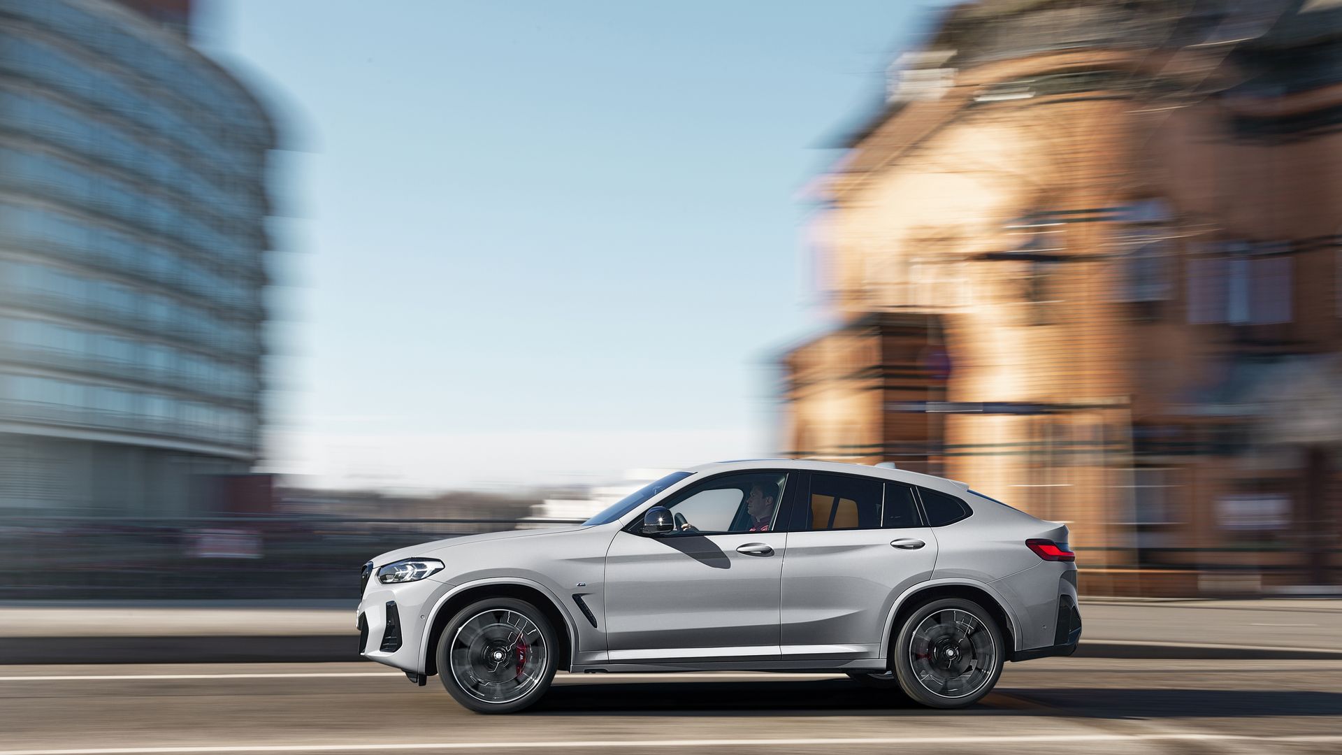 Silver SUV driving fast on a city street with blurred buildings in the background under a clear blue sky.