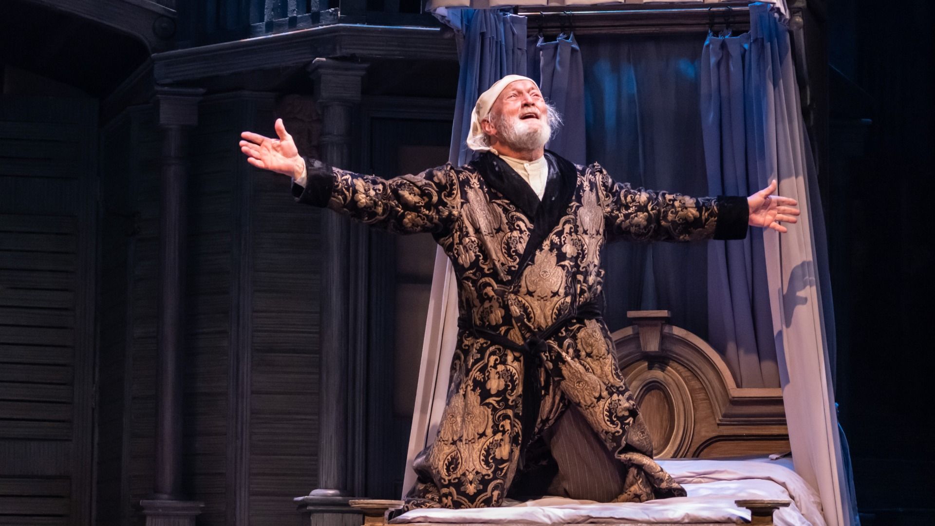 Elderly man in a patterned robe and nightcap kneels on a bed with arms outstretched, expressing joy or relief on a theatrical stage with blue curtains and classical backdrop.