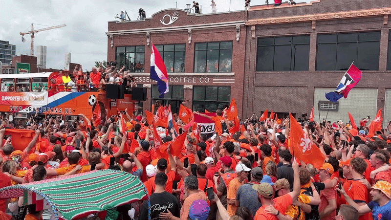 A crowded street filled with fans in orange, waving orange flags and a Dutch flag from a parade bus. People on a brick building overlook the scene as a festive Netherlands celebration unfolds.