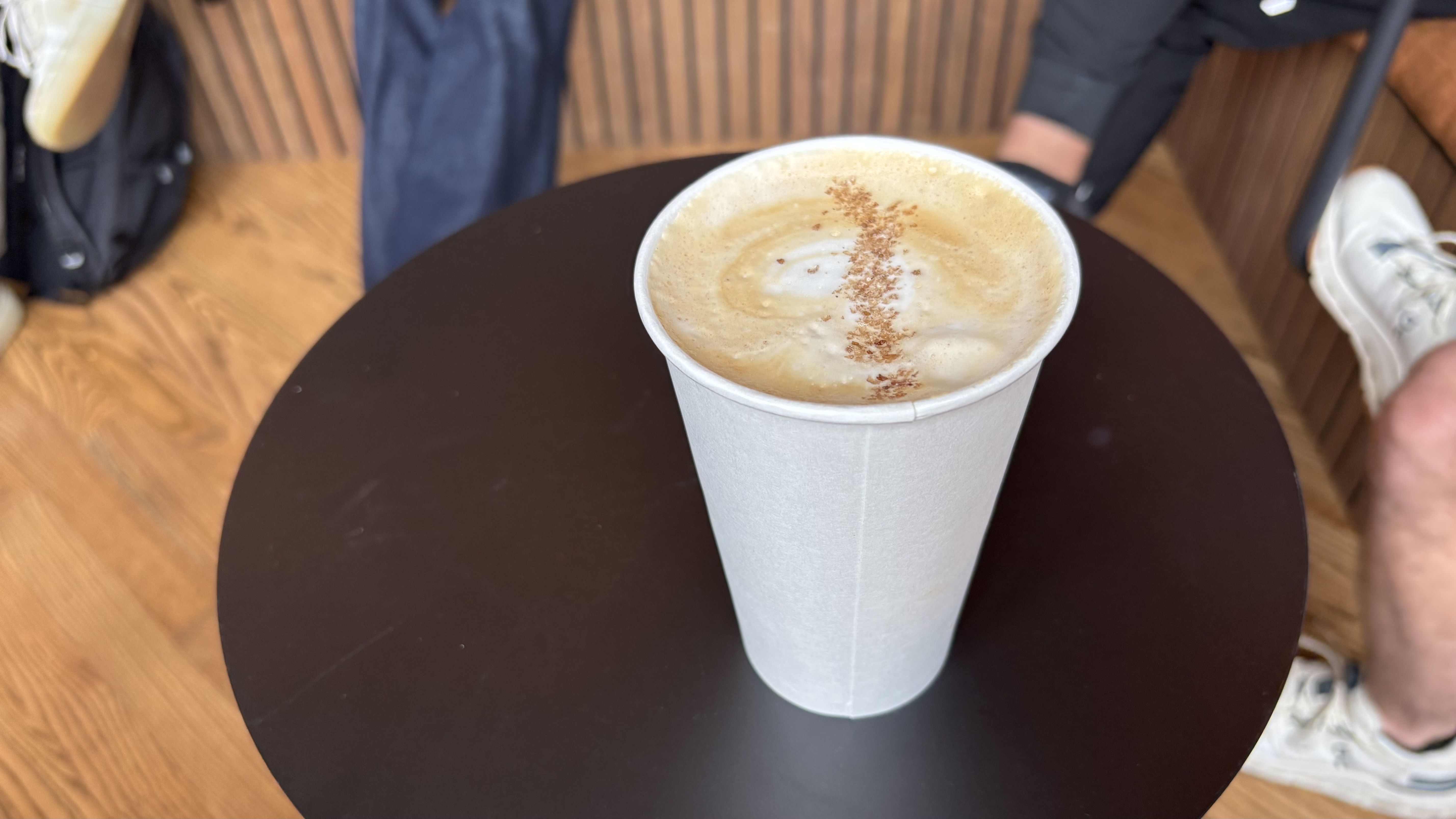 White paper cup filled with frothy coffee topped with a line of cinnamon on a dark round table, with wooden floor and legs of people wearing casual shoes in the background.