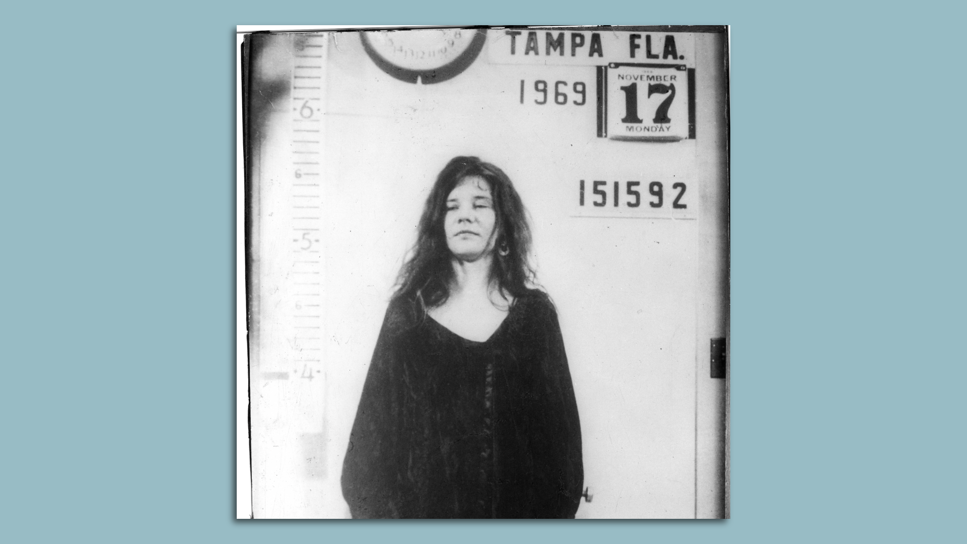 A black-and-white image of a woman with long dark hair in a dark-colored tunic. Behind her, writing on the wall says "TAMPA FLA. 1969 Nov. 17 151592."