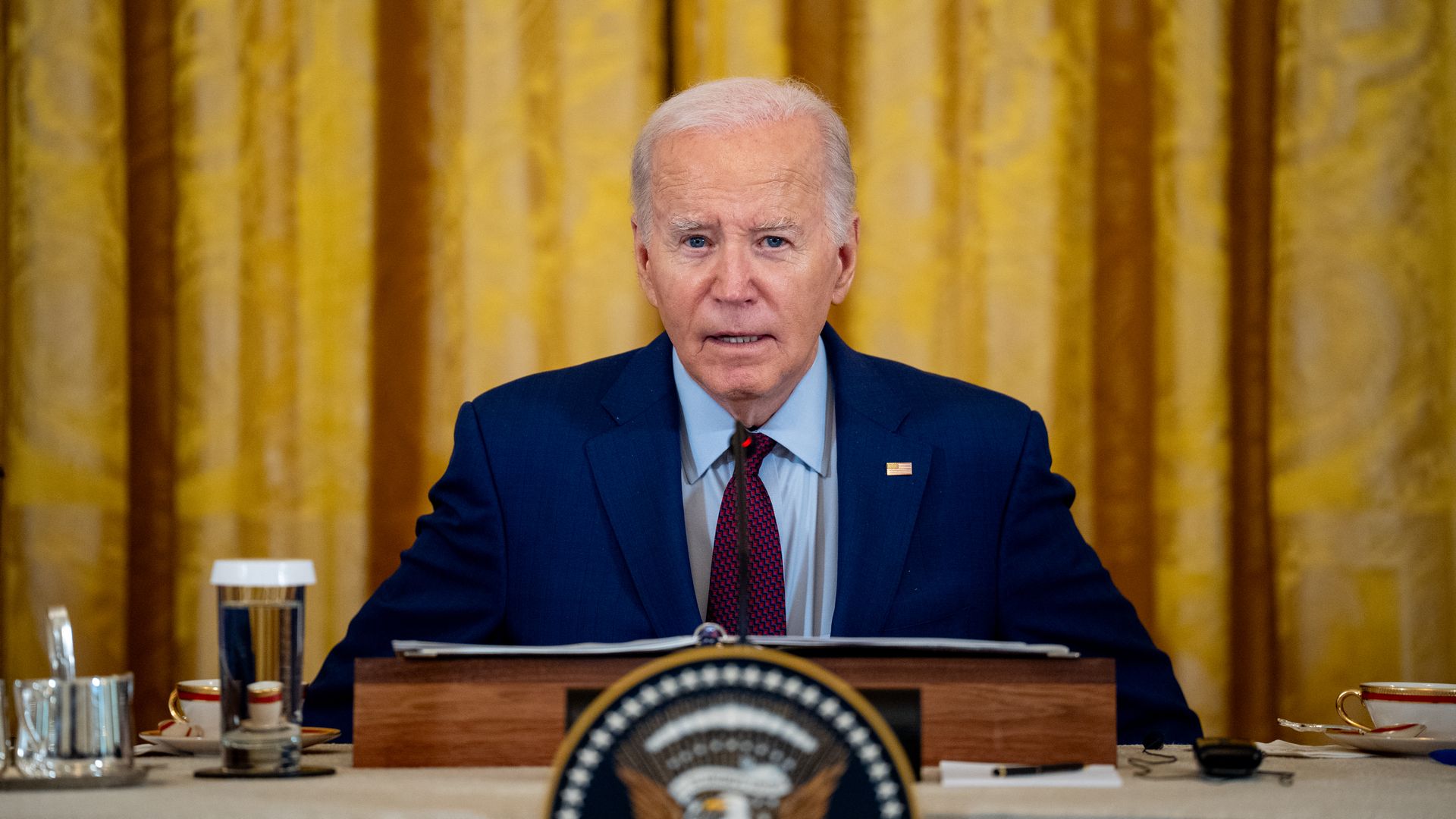 President Biden, wearing a blue suit, light blue shirt and maroon tie, speaking at a podium on a table in front of a gold-colored curtain.