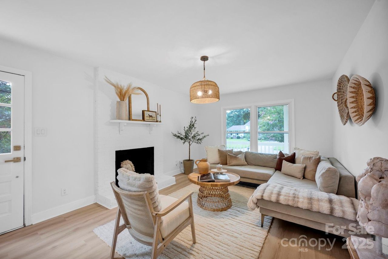 Bright living room with white walls, a large beige sectional sofa, wooden coffee table, woven hanging light, armchair with cushion, decorative fireplace, and two windows showing a green yard.