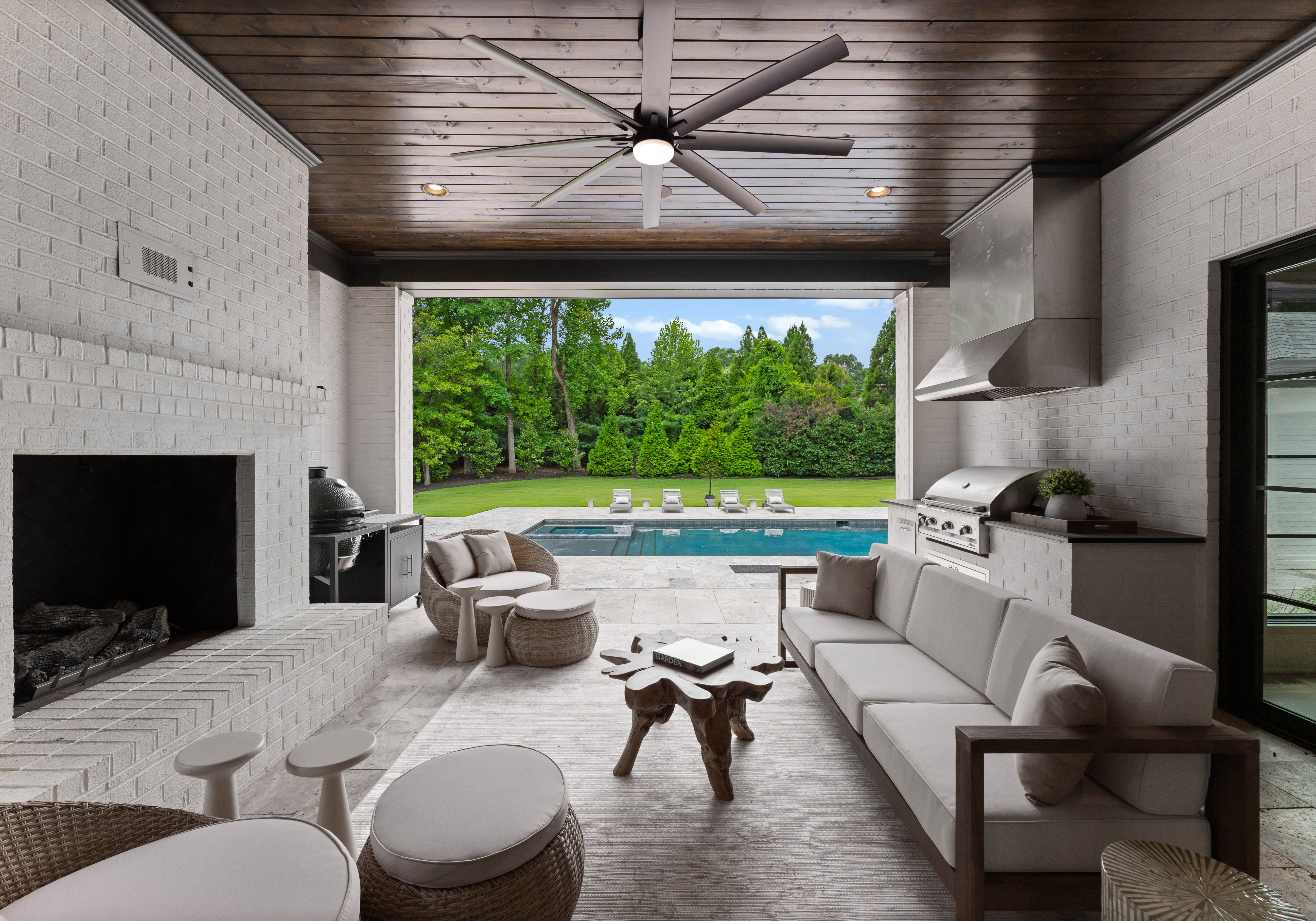Covered patio with white brick walls, wooden ceiling, large fan, and modern outdoor furniture overlooking a pool and green lawn with lounge chairs and trees in the background.