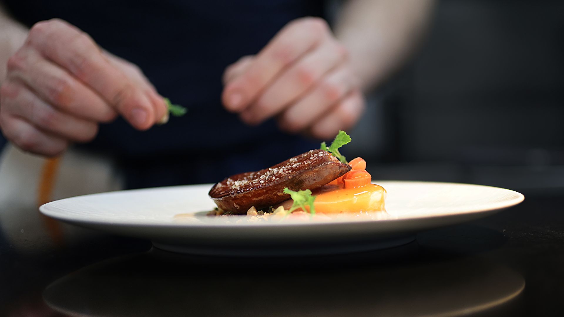 Close-up of a plated dish featuring a seared meat slice on orange sauce with greens; a chef garnishes the plate in the blurred background.