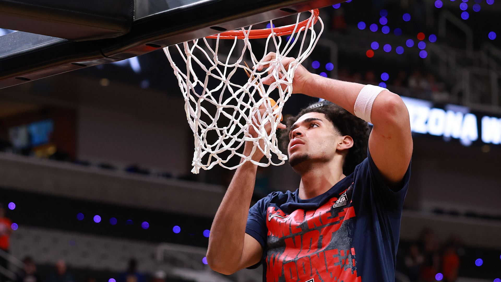 A young man in a blue t-shirt with red lettering and a University of Arizona cuts off part of a basketball net in a crowded arena.