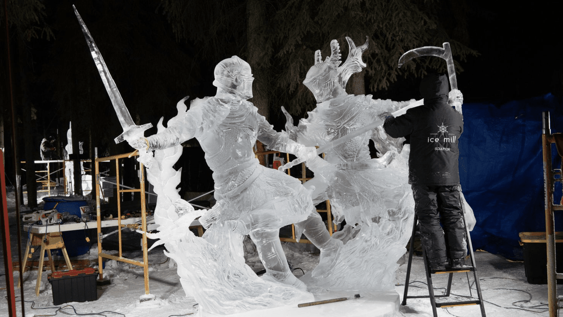 An artist wearing a black jacket labeled "ice mill" sculpts a detailed ice statue of two armored knights in combat, one holding a sword and the other a scythe, in an outdoor snowy setting at night.