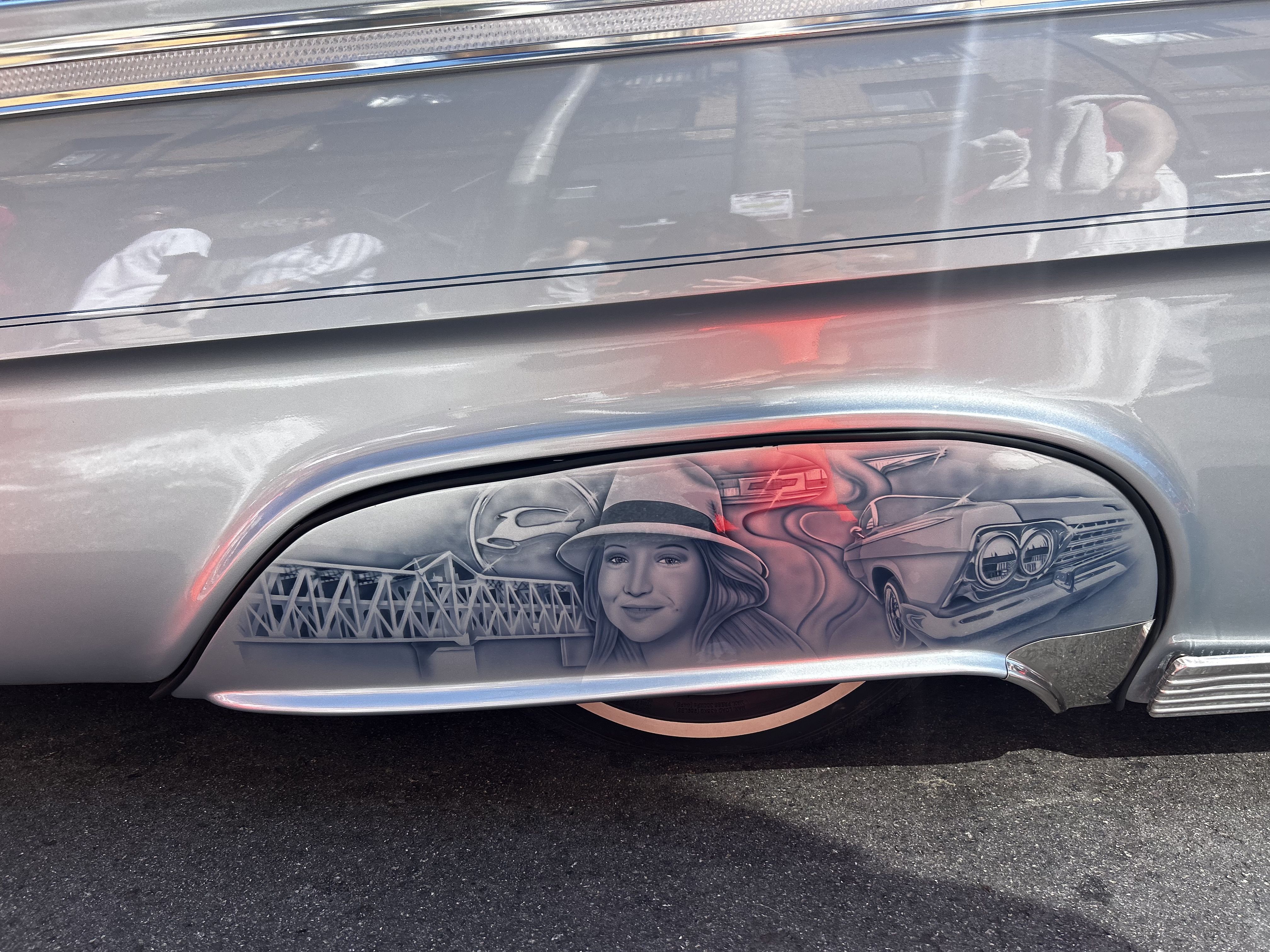 Photo of a silver lowrider car's side, which shows an intriate graphic of a woman, car and bridge
