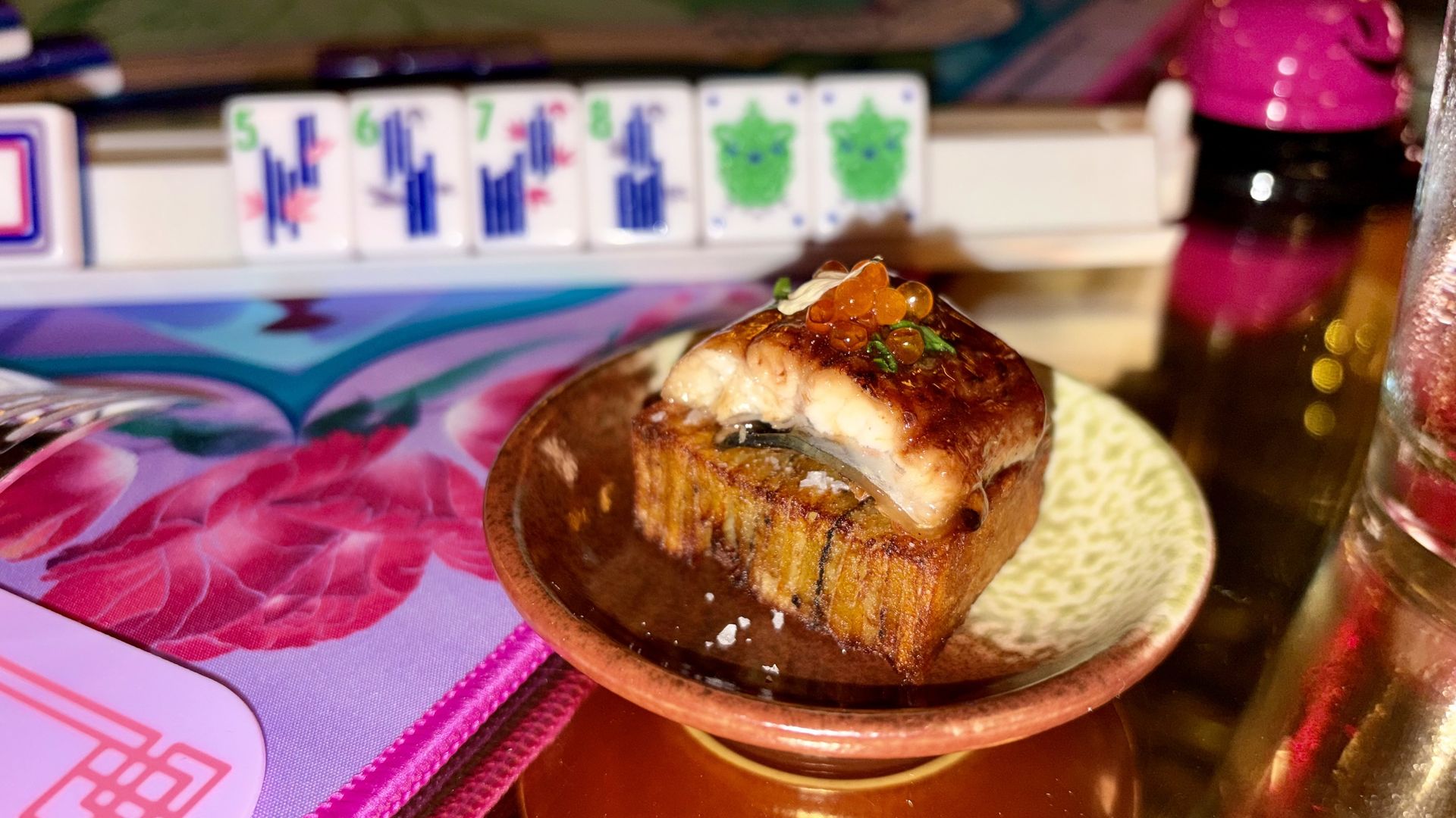 Orange fish roe sits on a tiny ceramic pedestal, on a colorful table. In the background, Mahjong tiles and a pink bottle are visible.