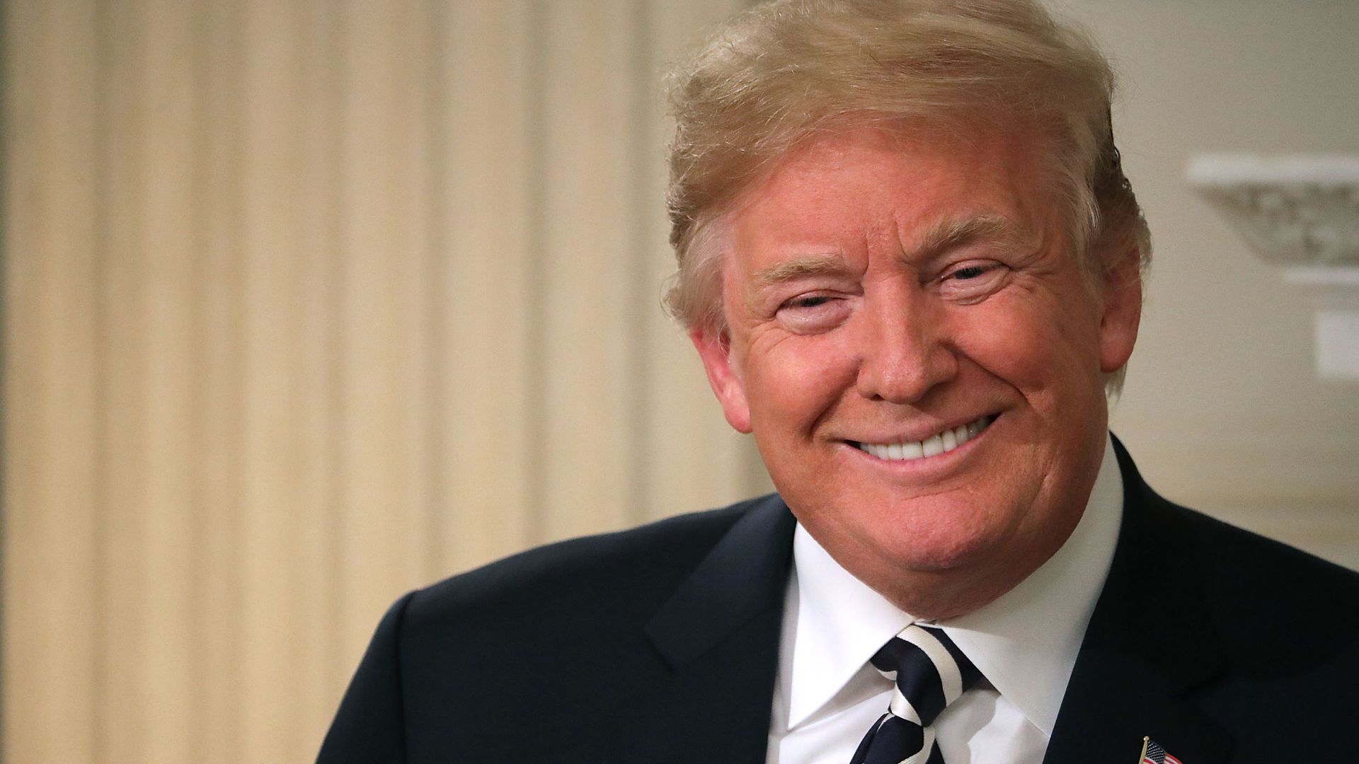 Trump smiles in a black suit before a white background.