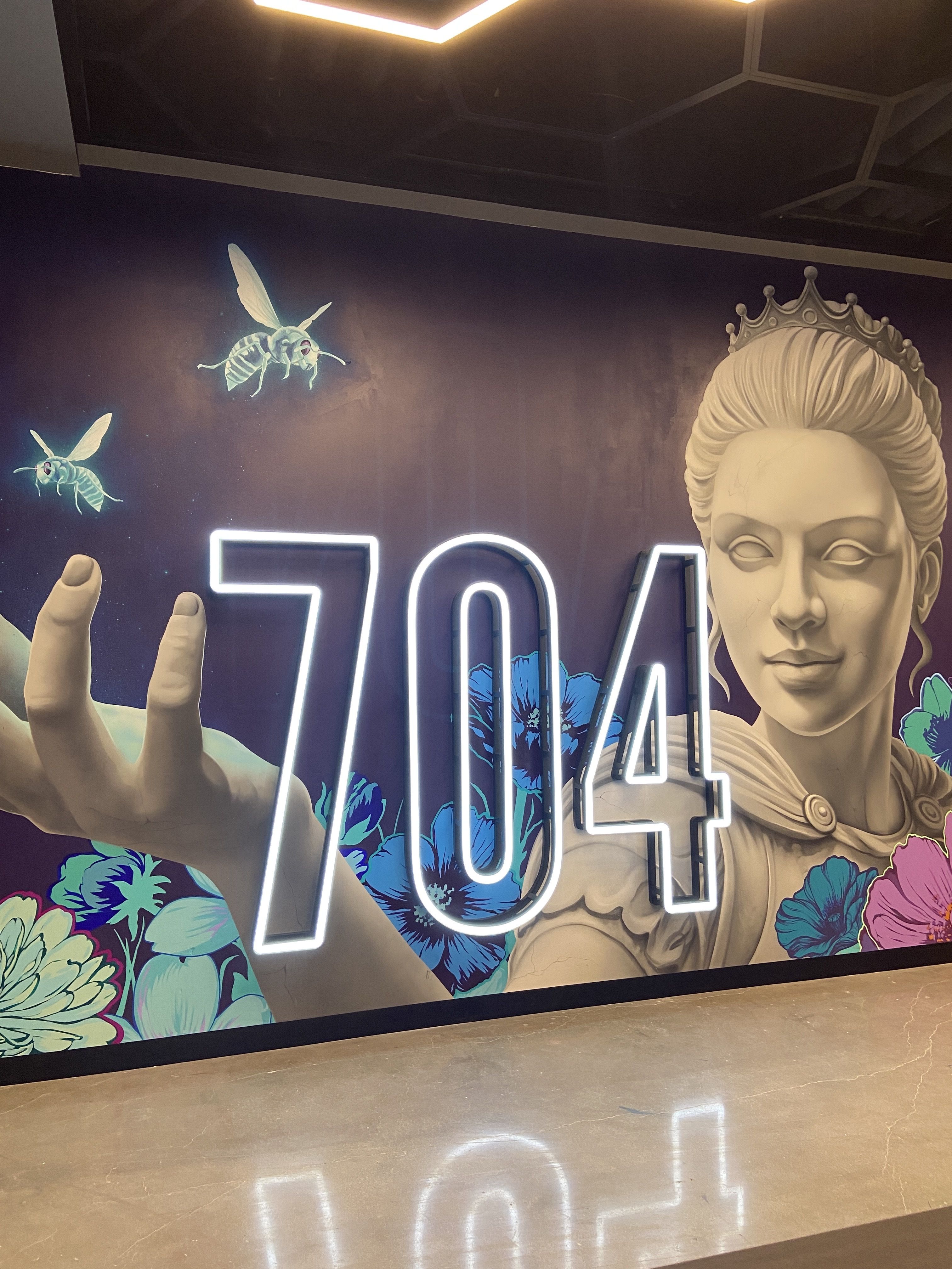 Mural with a large grayscale statue of a crowned woman extending her hand, surrounded by blue and purple flowers, two glowing bees, and bright neon numbers "704" on a dark wall.