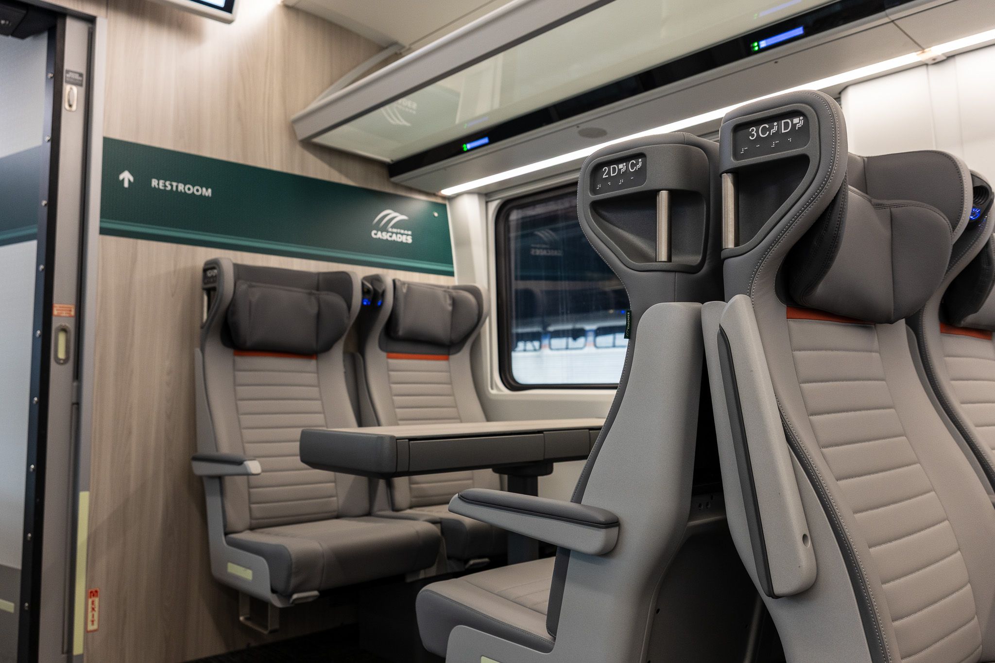 Interior of a modern train cabin with gray seats and armrests; a green sign reads "RESTROOM" with an arrow, and a "Cascades" logo is visible near the window.