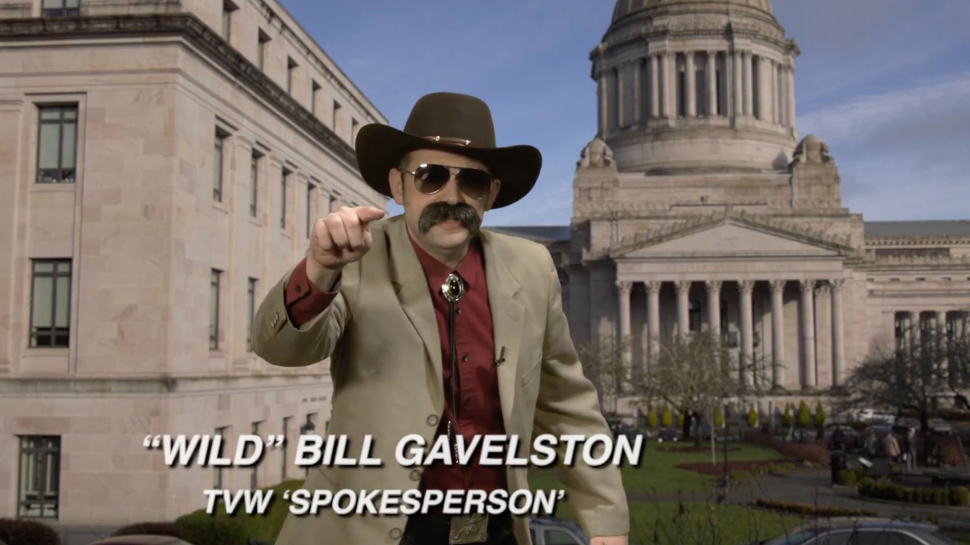 Man in a tan suit, red shirt, bolo tie, cowboy hat and sunglasses with a large mustache points at the camera. A domed capitol building stands behind; captions read "WILD BILL GAVELSTON" and TVW "SPOKESPERSON".