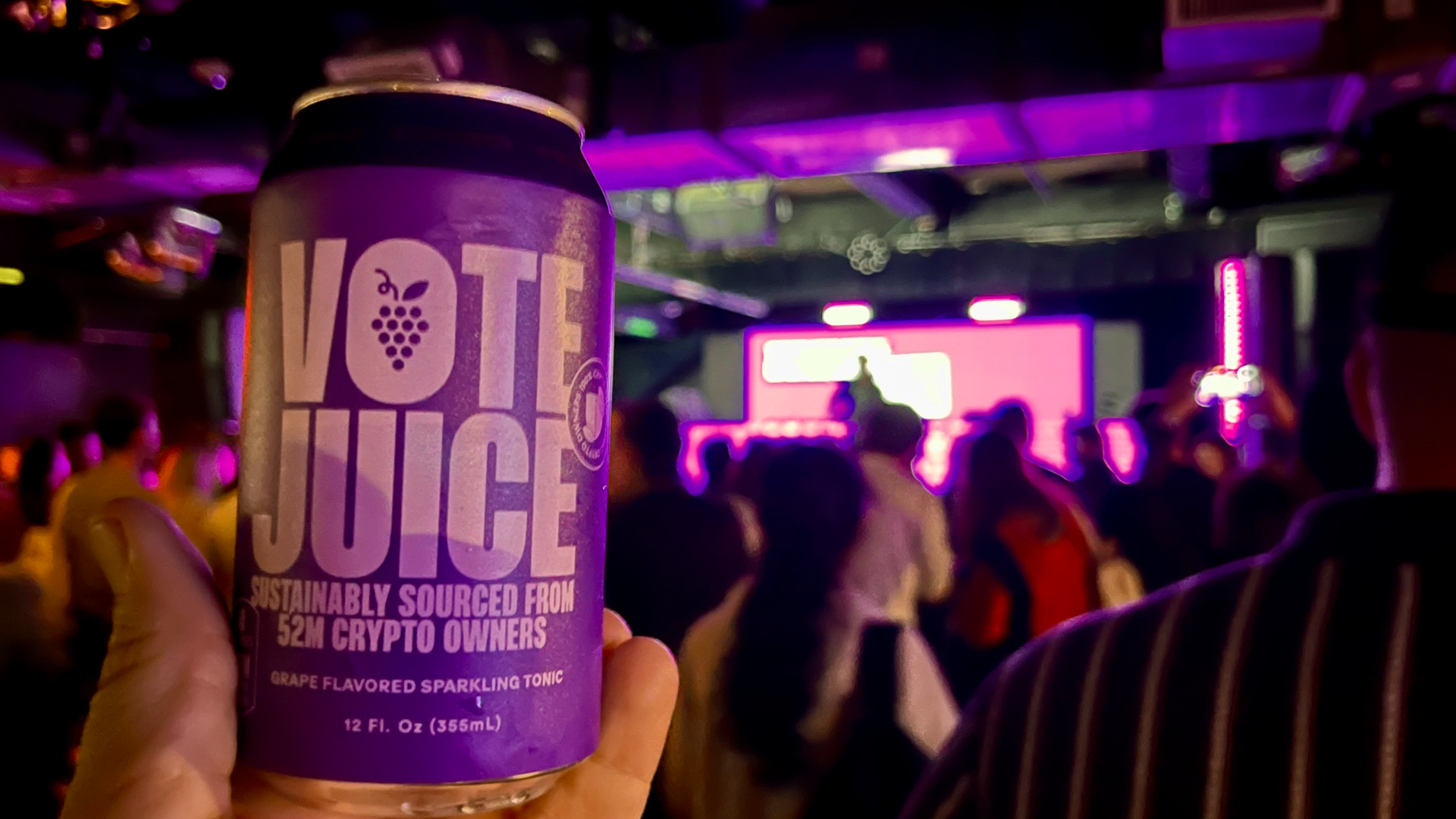 A can of "Vote Juice," largely in purple, in front of a bunch of people watching a DJ in a dark room.