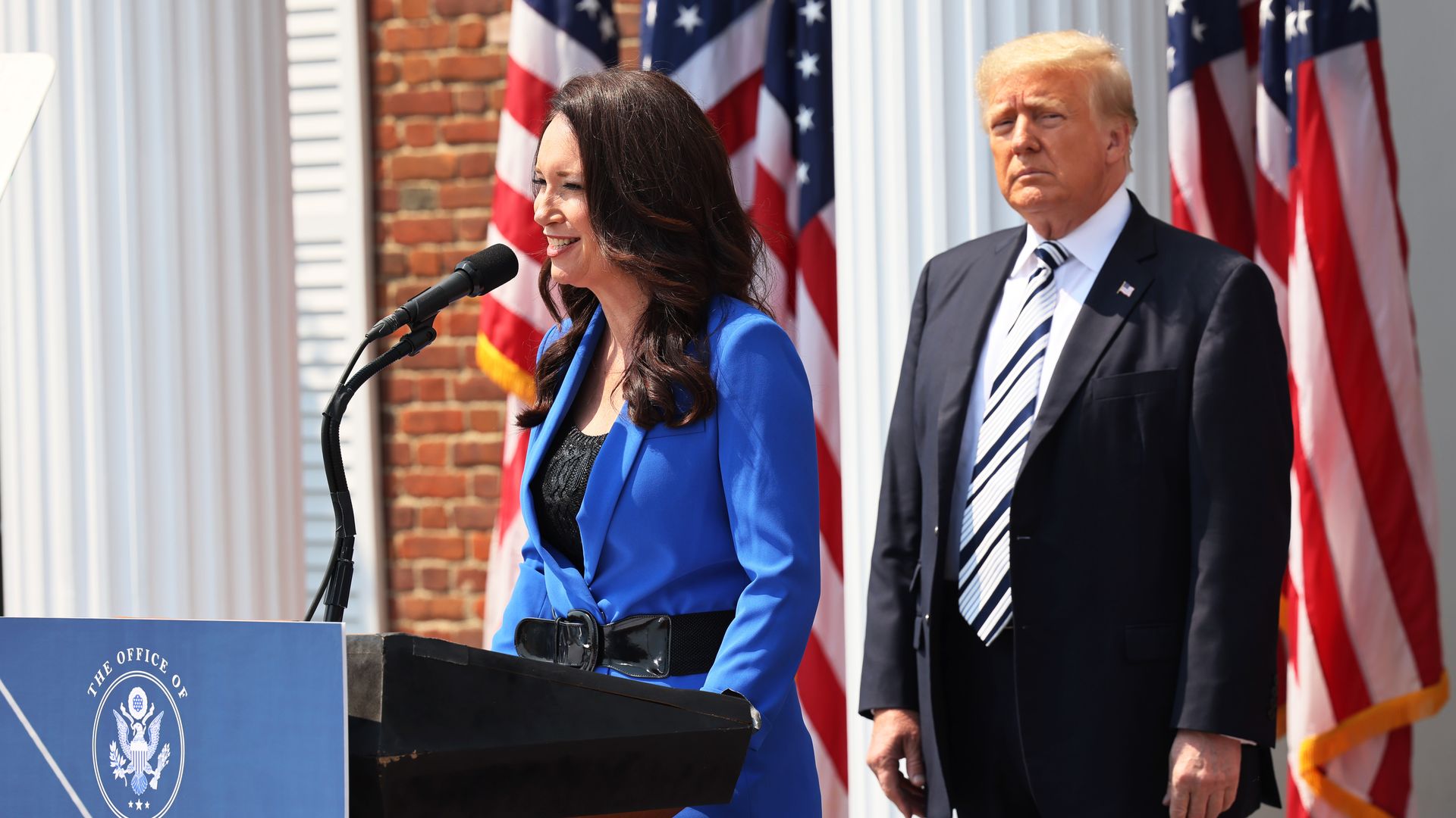 Brooke Rollins speaks at a podium while Donald Trump stands behind her.