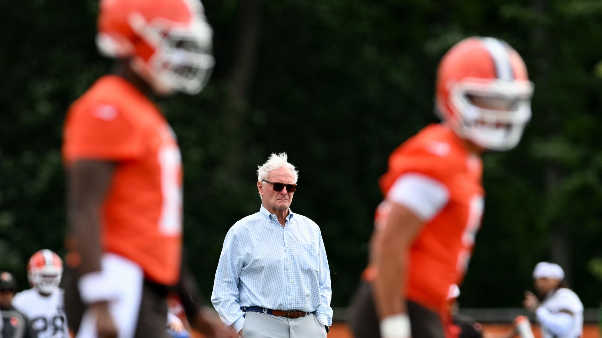 Browns owner Jimmy Haslam, wearing sunglasses and light blue checkered shirt stands with hands in pockets on a football field, flanked by two blurred players in orange jerseys and helmets.