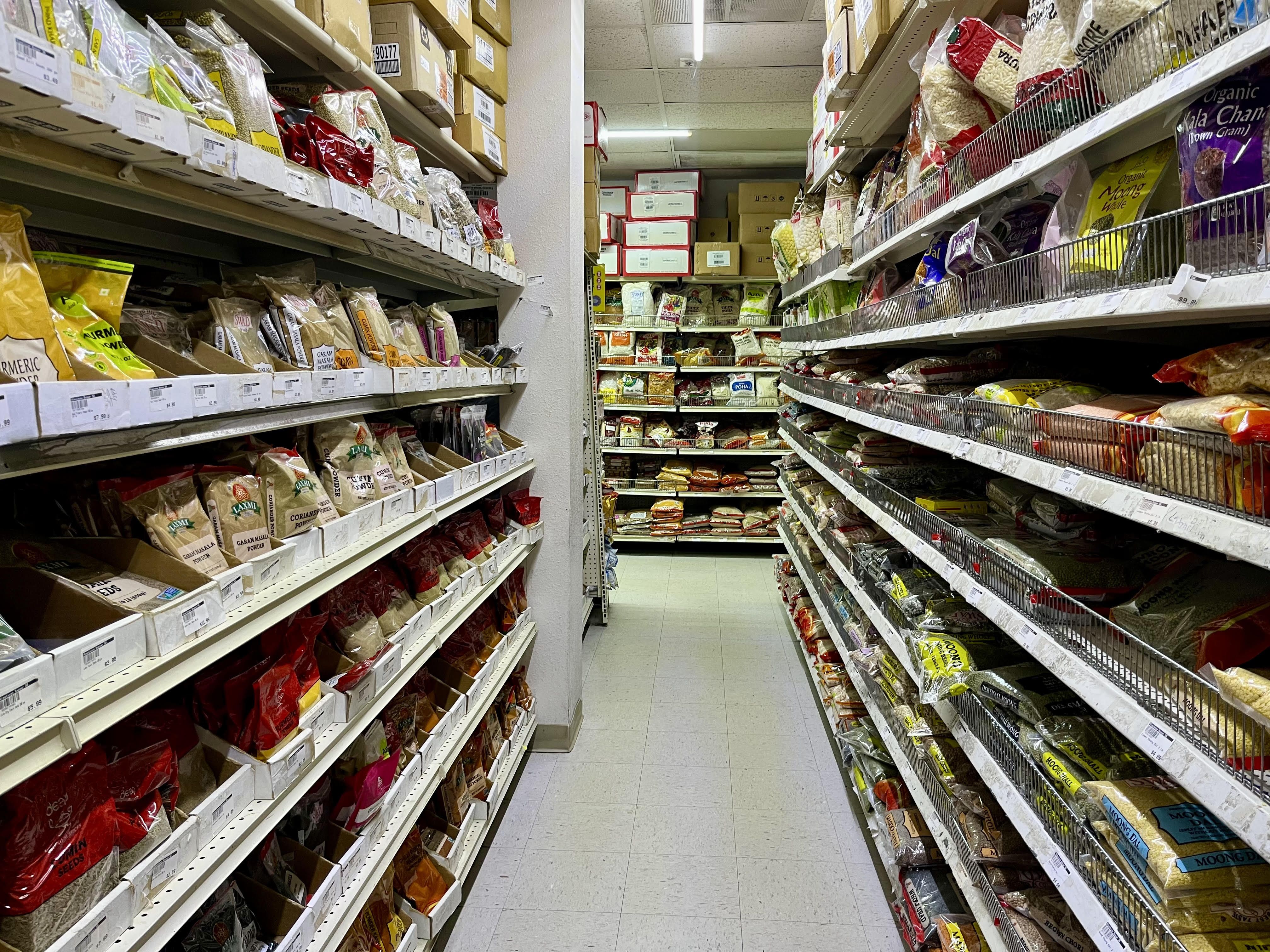 A well-lit grocery aisle at the Himalayan Bazar in San Antonio with shelves stocked with various packaged spices, grains, and dry goods in clear and colorful bags, including turmeric, coriander, and dals.