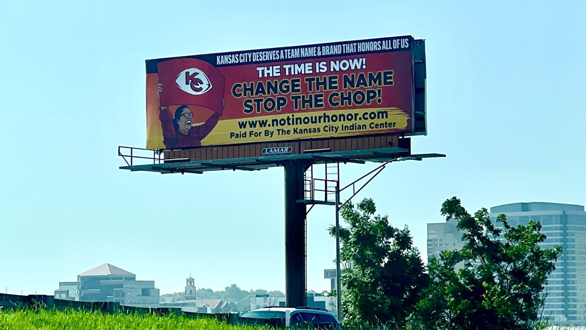 Colorful roadside billboard on a tall pole with bold text reading THE TIME IS NOW! CHANGE THE NAME STOP THE CHOP, plus a cheering figure holding a KC logo and the site notinourhonor.com.