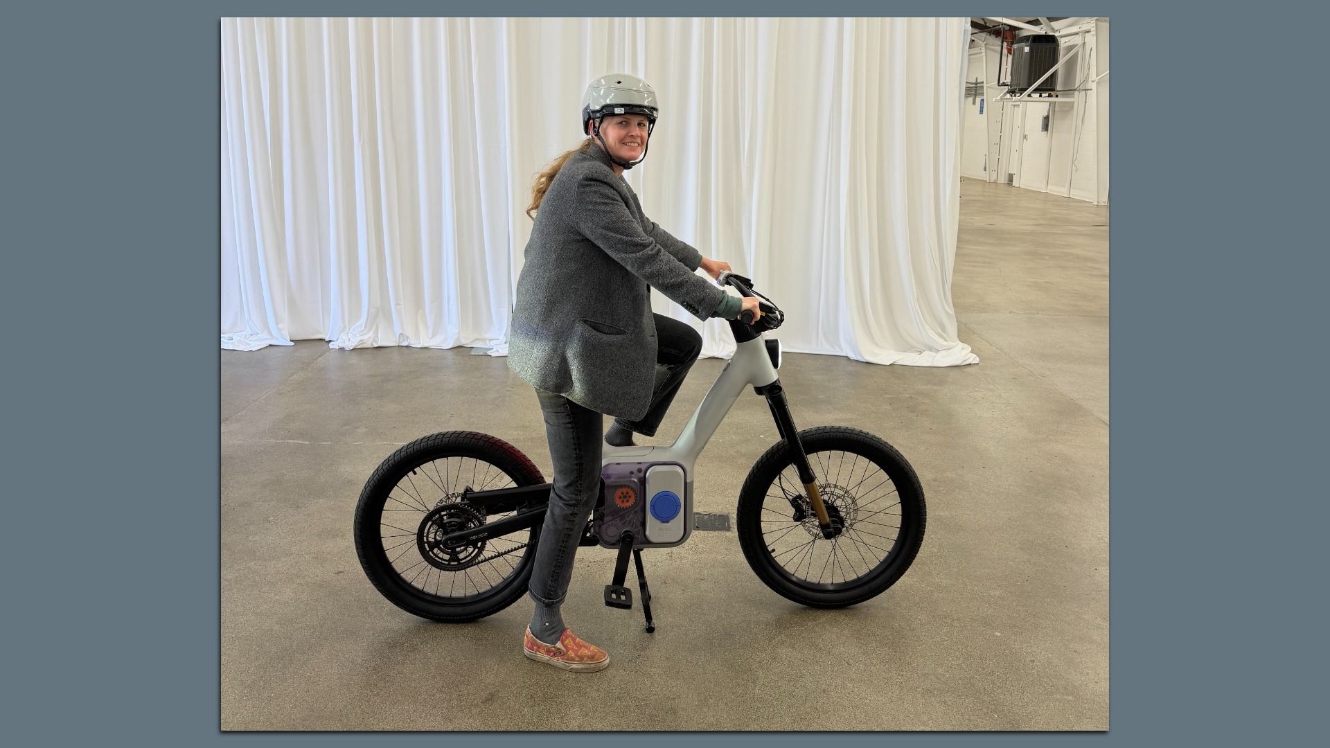 Person wearing a gray helmet, gray jacket, and black pants sitting on a modern gray and black electric bike indoors with white curtains in the background.