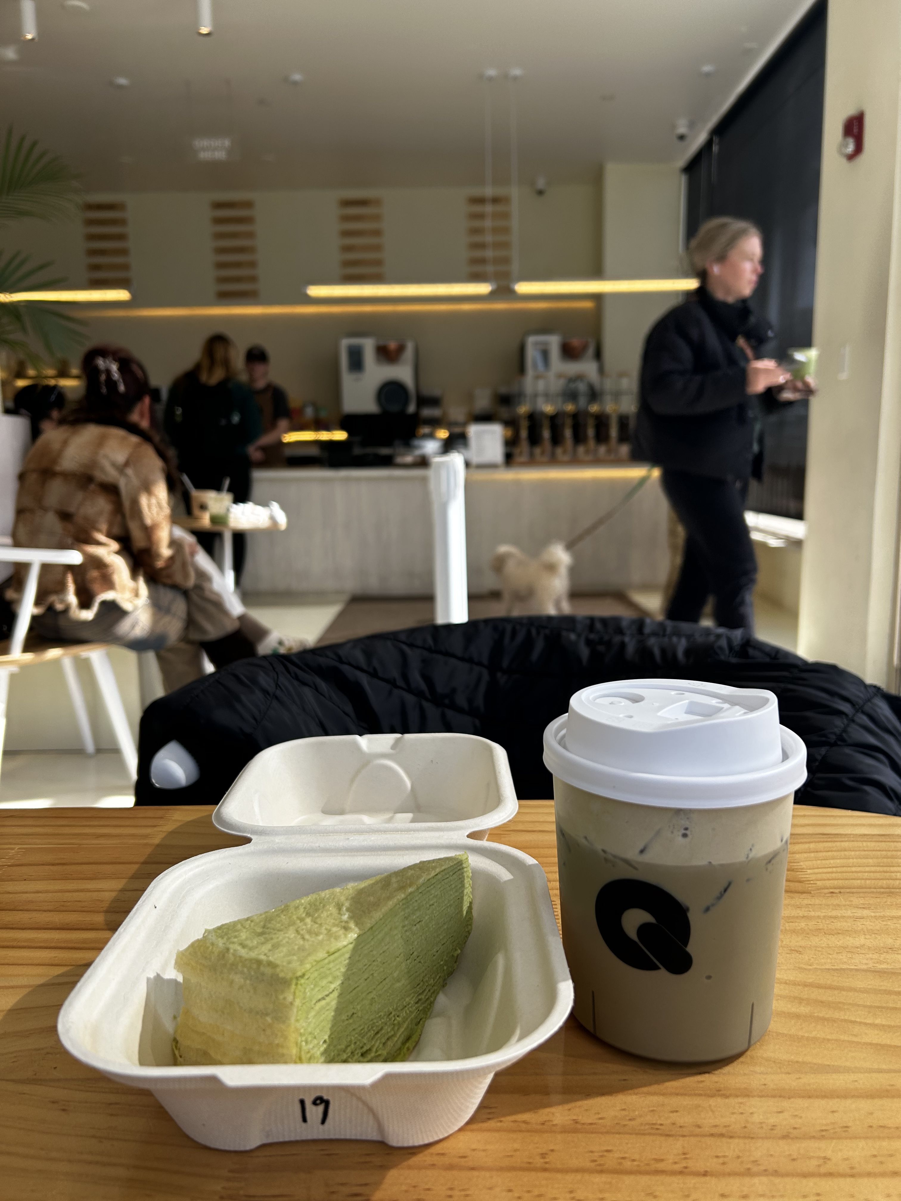 Cafe scene: green tea cake in a white takeout tray and a beige iced drink on a wooden table. In the background, people and a person with a leash dog near the counter.