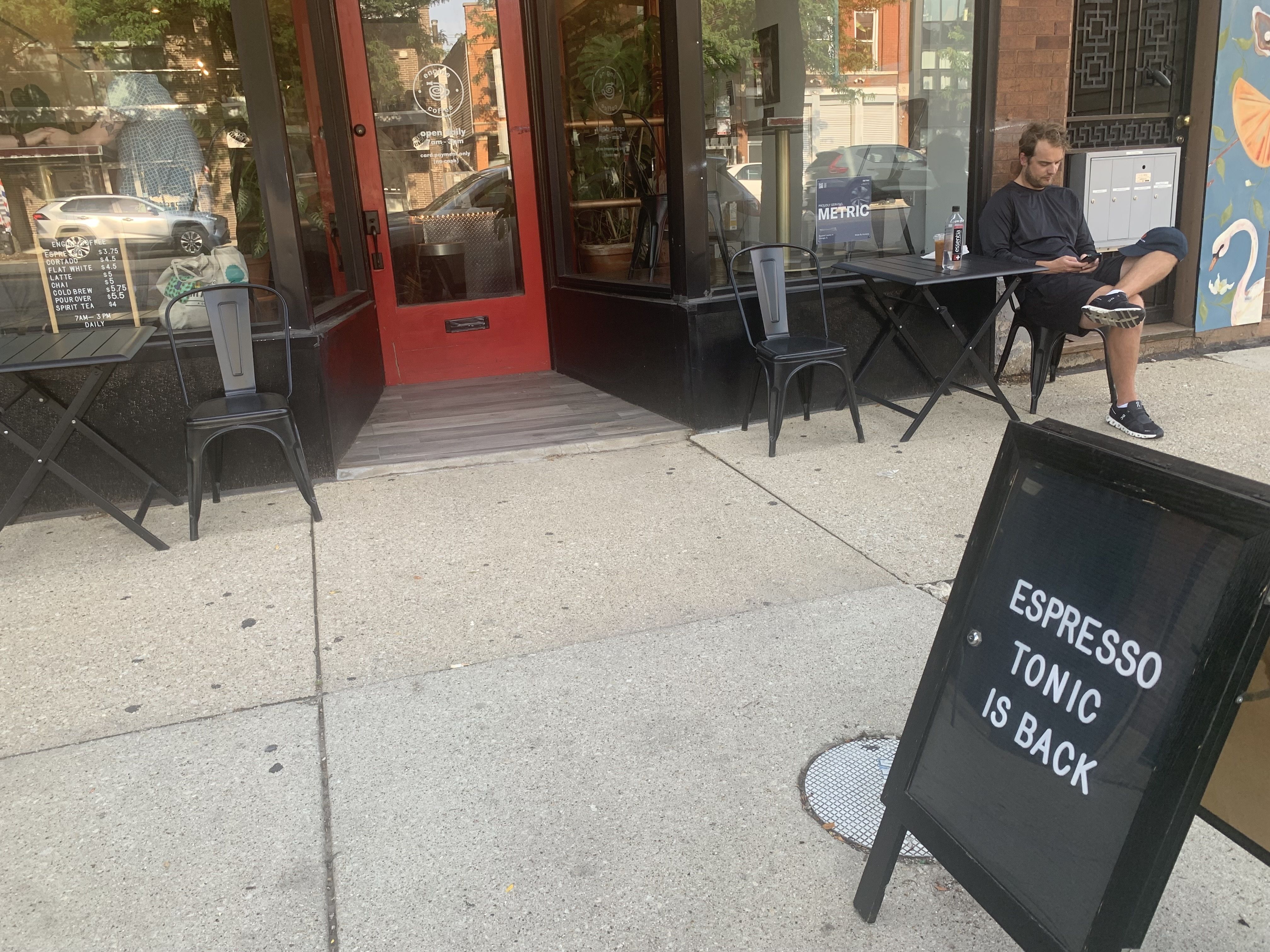 Outdoor seating at a coffee shop with a bright red door, black tables and chairs, and a black sandwich board sign reading "ESPRESSO TONIC IS BACK." A man sits at a table using his phone.