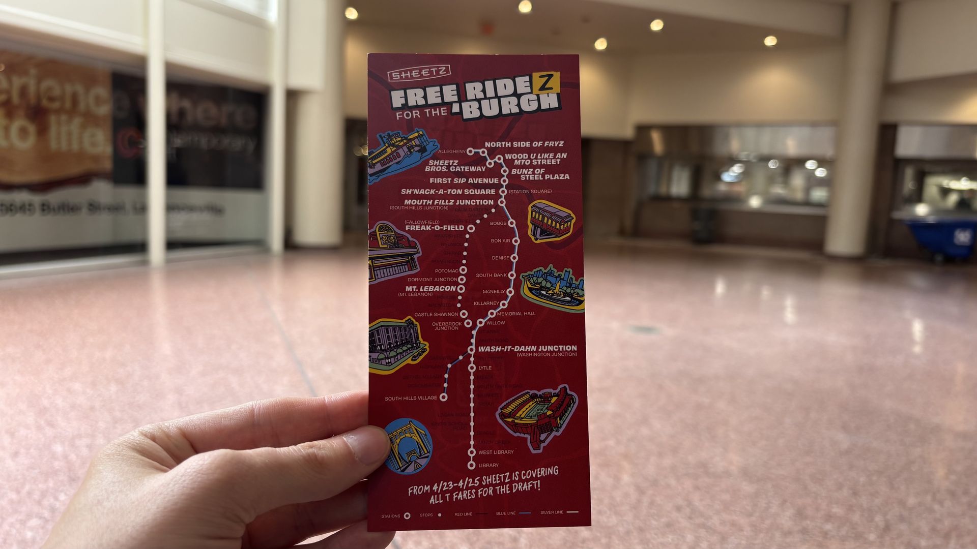 A hand holds a red brochure titled "Free Ride for the Burgh" displaying a transit map with cartoon trains, set against a blurred mall interior.