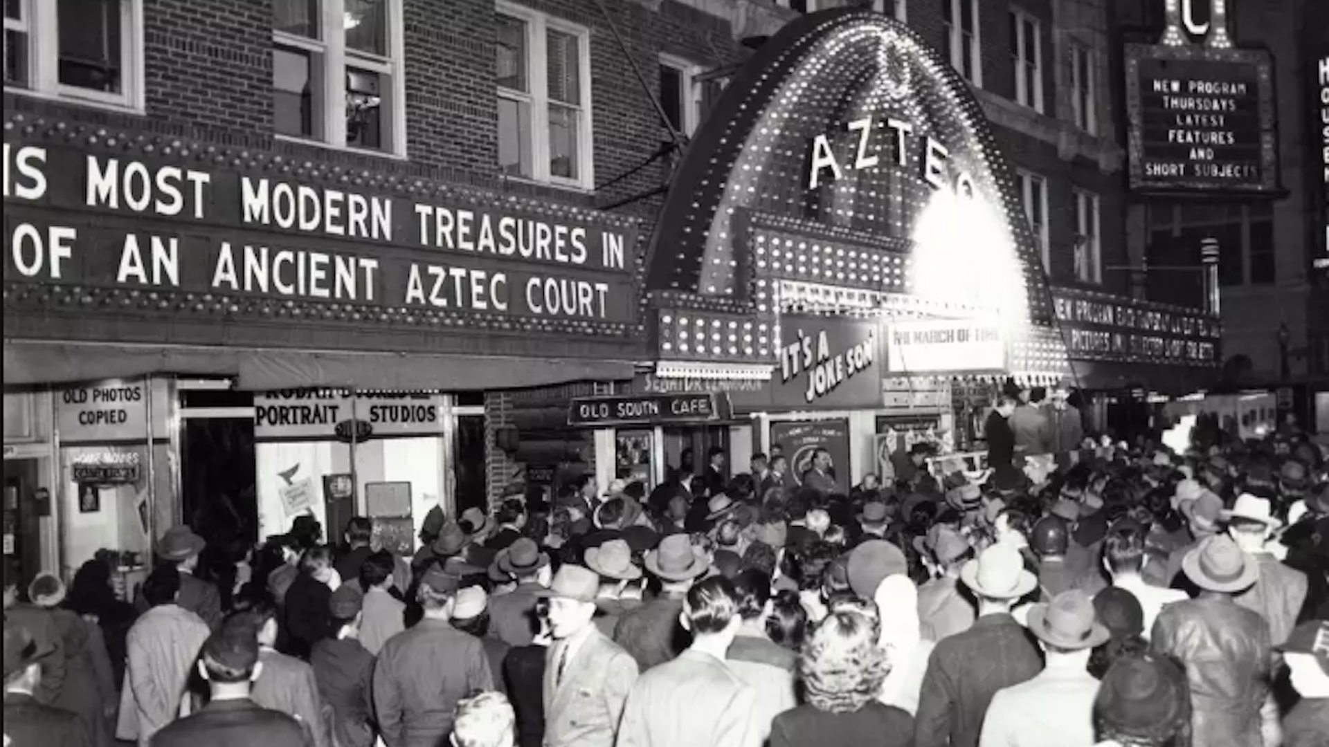 Black-and-white street scene outside a vintage theater with a bright 'AZTEC' marquee; a dense crowd in 1940s attire fills the sidewalk as storefronts advertise portrait studios and old photos.