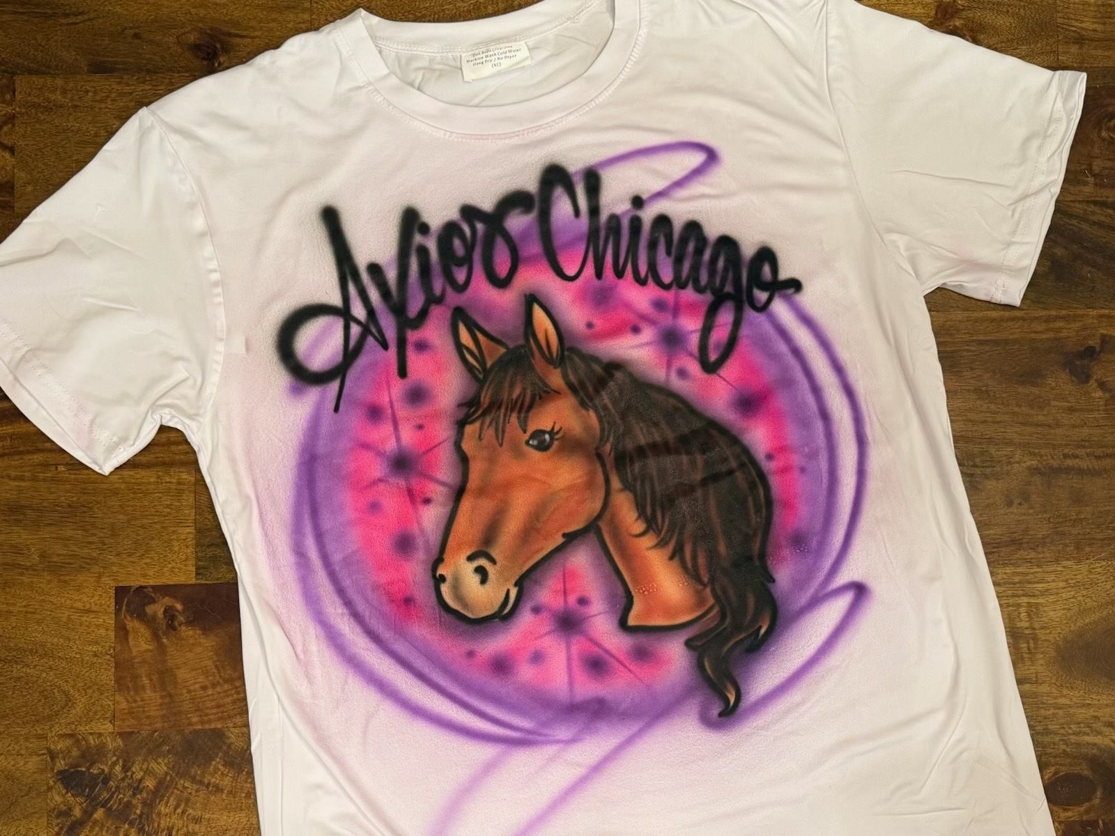 White T-shirt with graffiti-style design: a brown horse head with a dark mane in the center, surrounded by pink and purple spray circles, and black cursive lettering across the top.