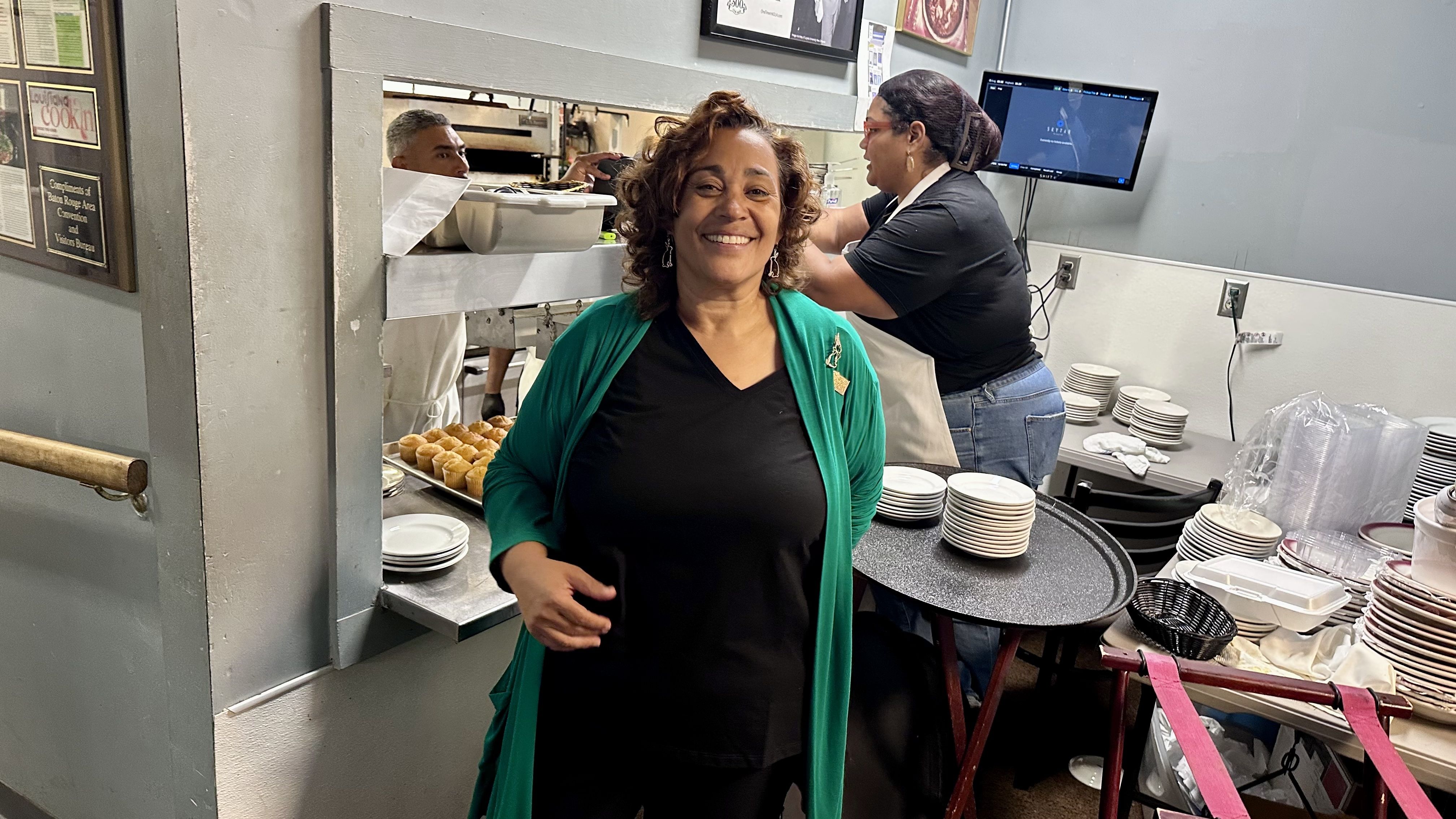 Smiling woman in a green cardigan stands in a busy kitchen, with muffins on a tray behind her and a line cook at the pass. Stacks of plates and a monitor are visible in the background.