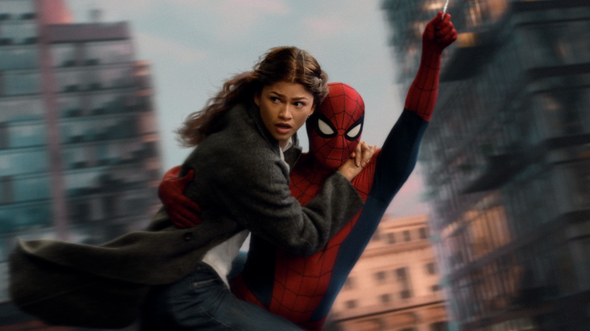 A woman in a gray coat clings to Spider-Man as they swing over a city, wind whipping her hair. Spider-Man’s red-and-blue suit and mask loom against blurred buildings.