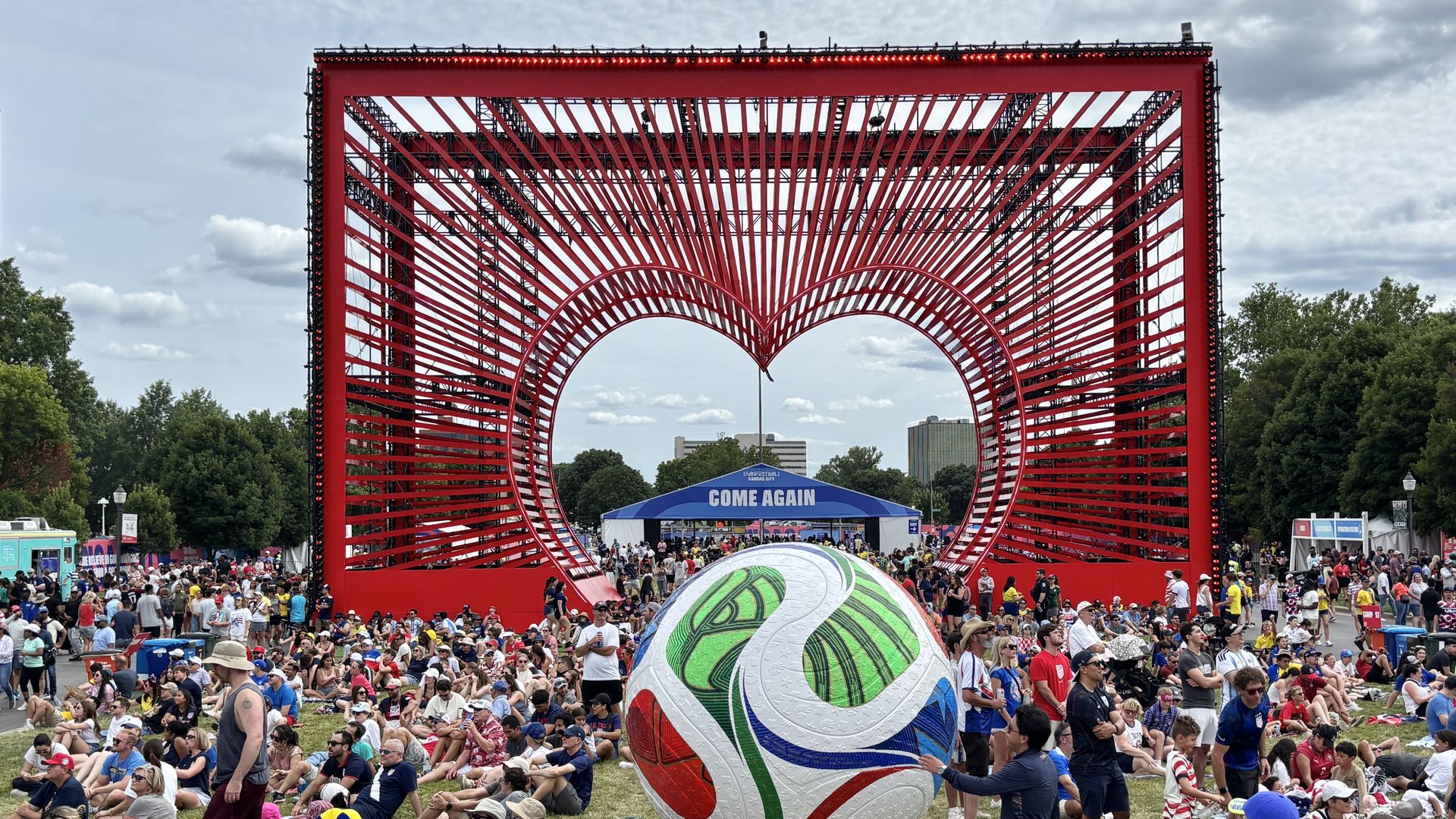 Crowded outdoor festival beneath a giant red geometric frame sculpture with curved stripes; a blue tent reads "COME AGAIN" in background, a colorful ball in foreground, trees and cloudy sky.