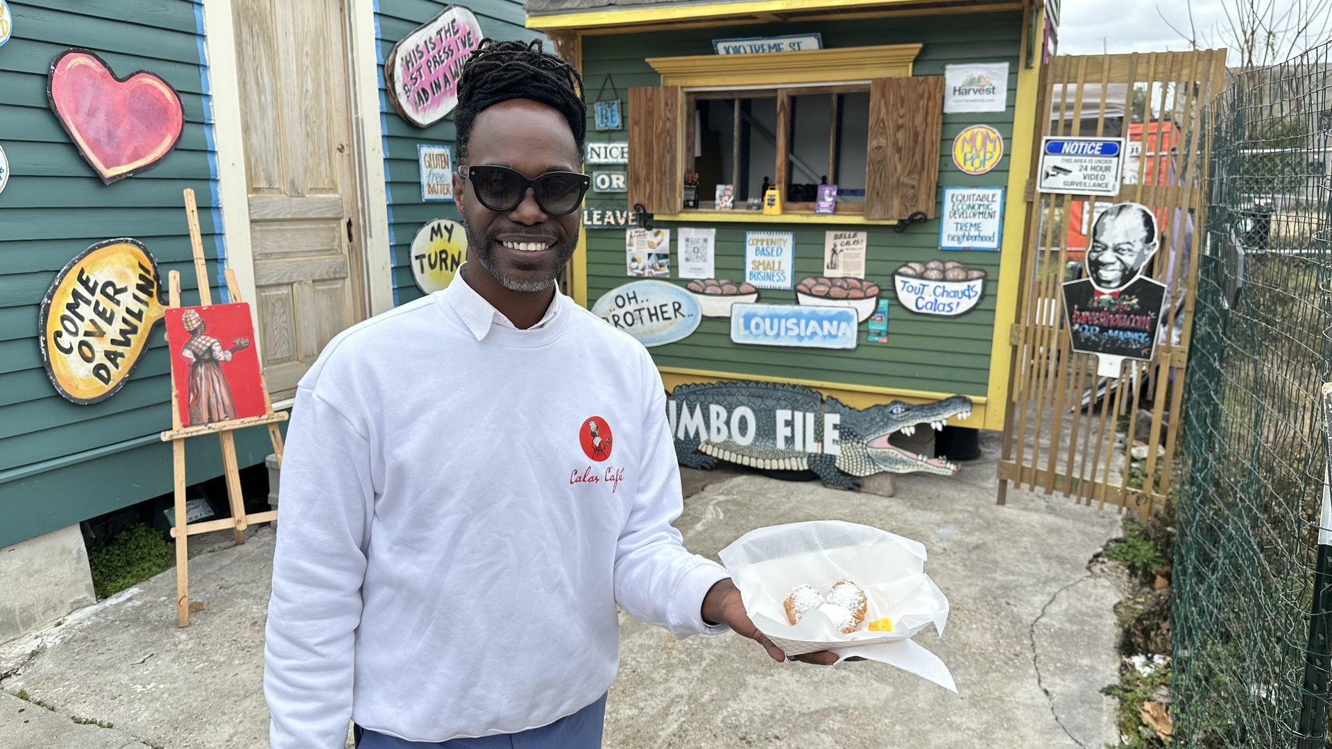 Brandan Pellerin holds up a paper tray of calas, smiling as he stands in front of The Calas Cafe. The food cart has brightly colored signs advertising the food and its history.