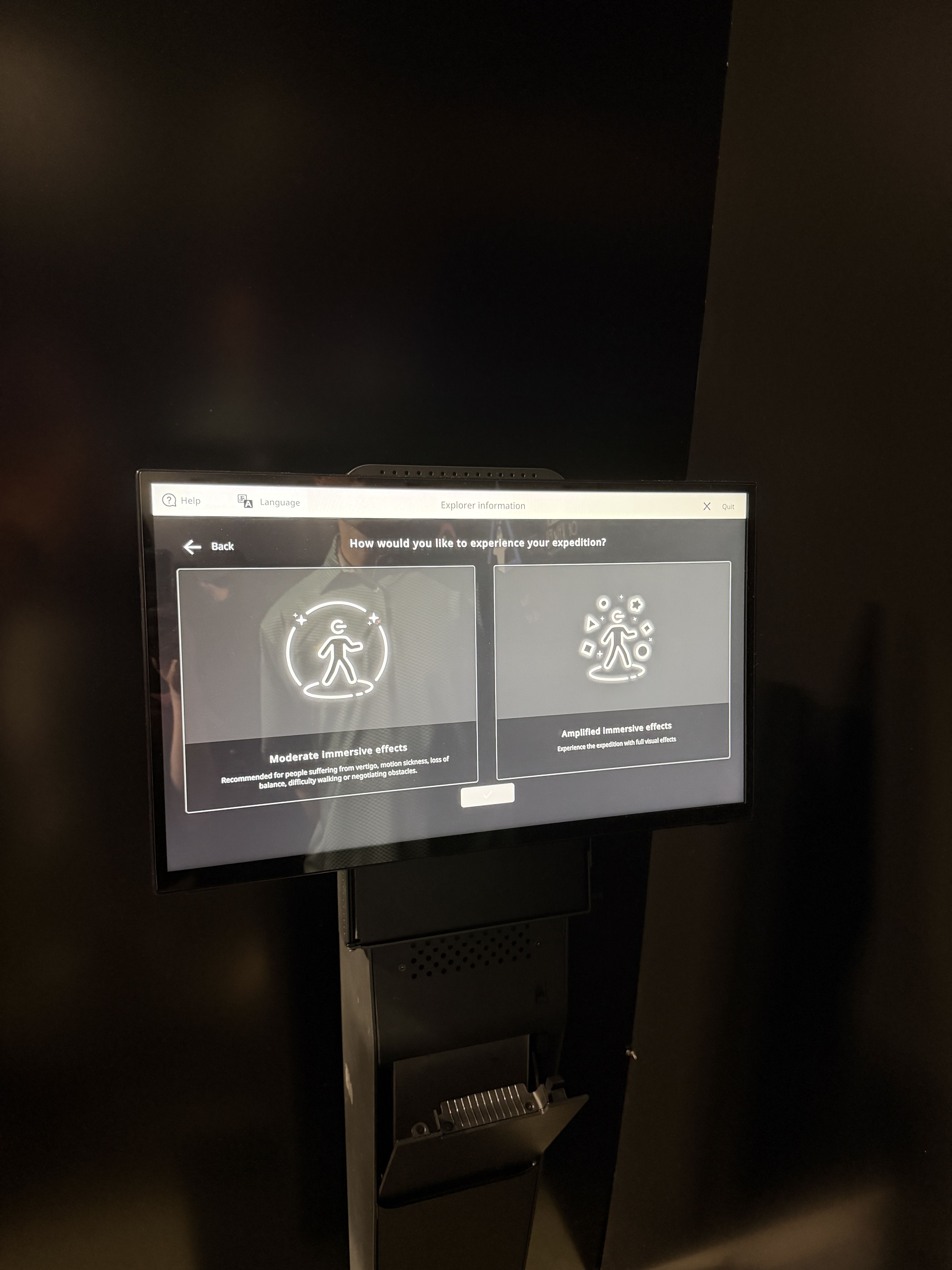 Dimly lit wall with a bottom-mounted touchscreen kiosk. The screen shows a dark UI with two panels, each featuring a white outline icon of a person and exploration motifs.