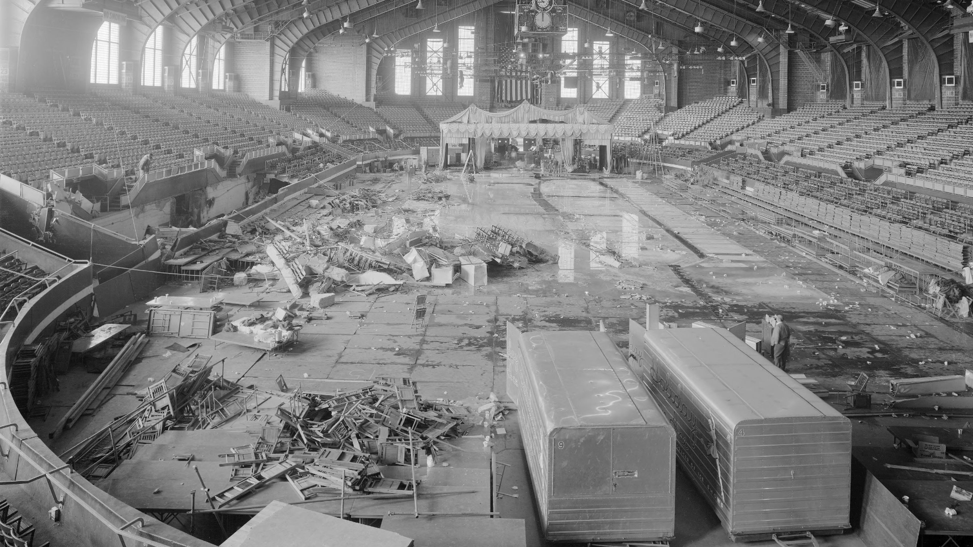 Black-and-white photo of a vast indoor arena under renovation. An arched steel roof, empty seating on both sides, scattered debris across the floor, and a white circus-style tent at center stage.