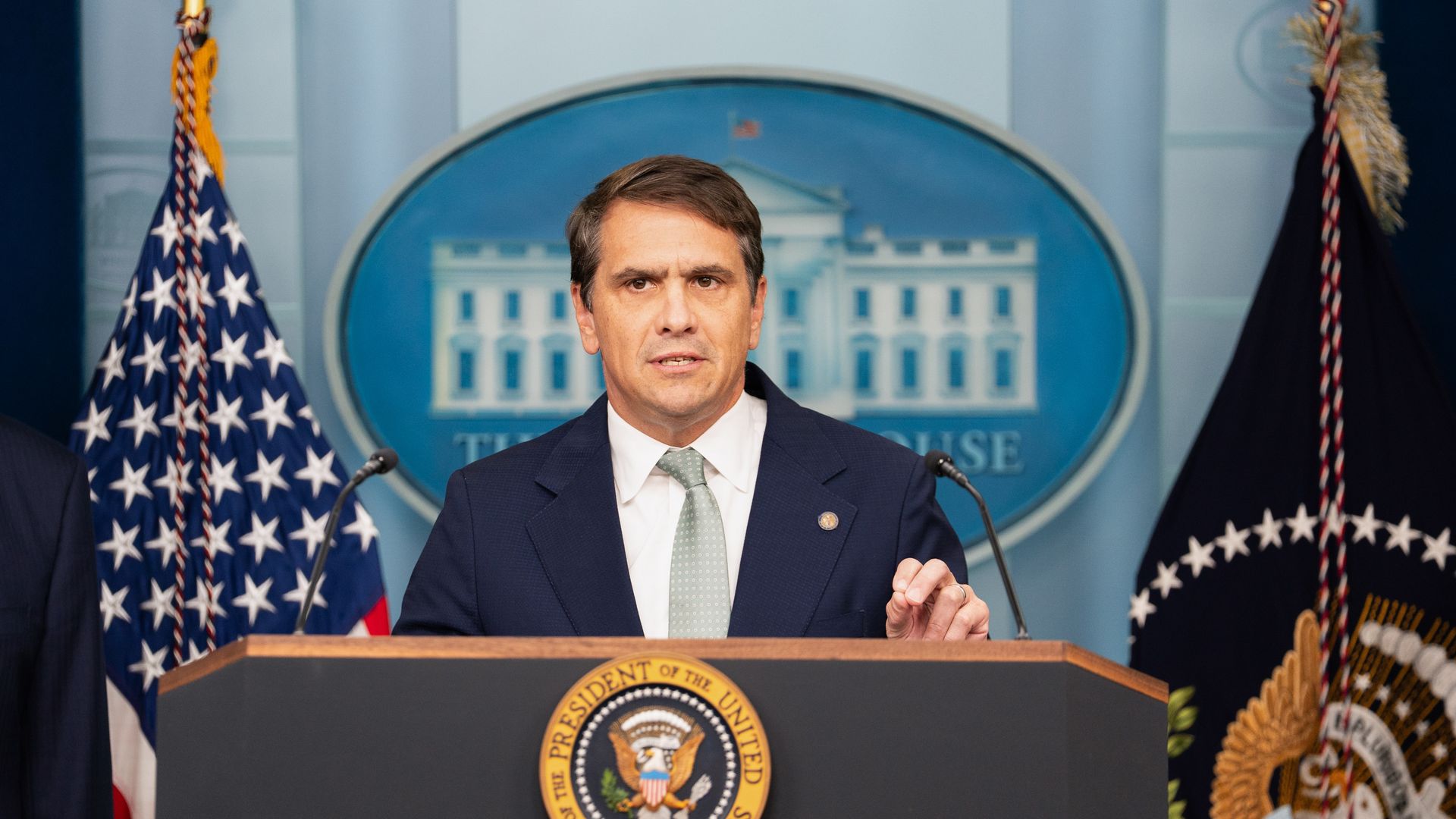 Todd Blanche, wearing a dark suit and a green tie — speaks from behind a podium with the White House seal behind him.