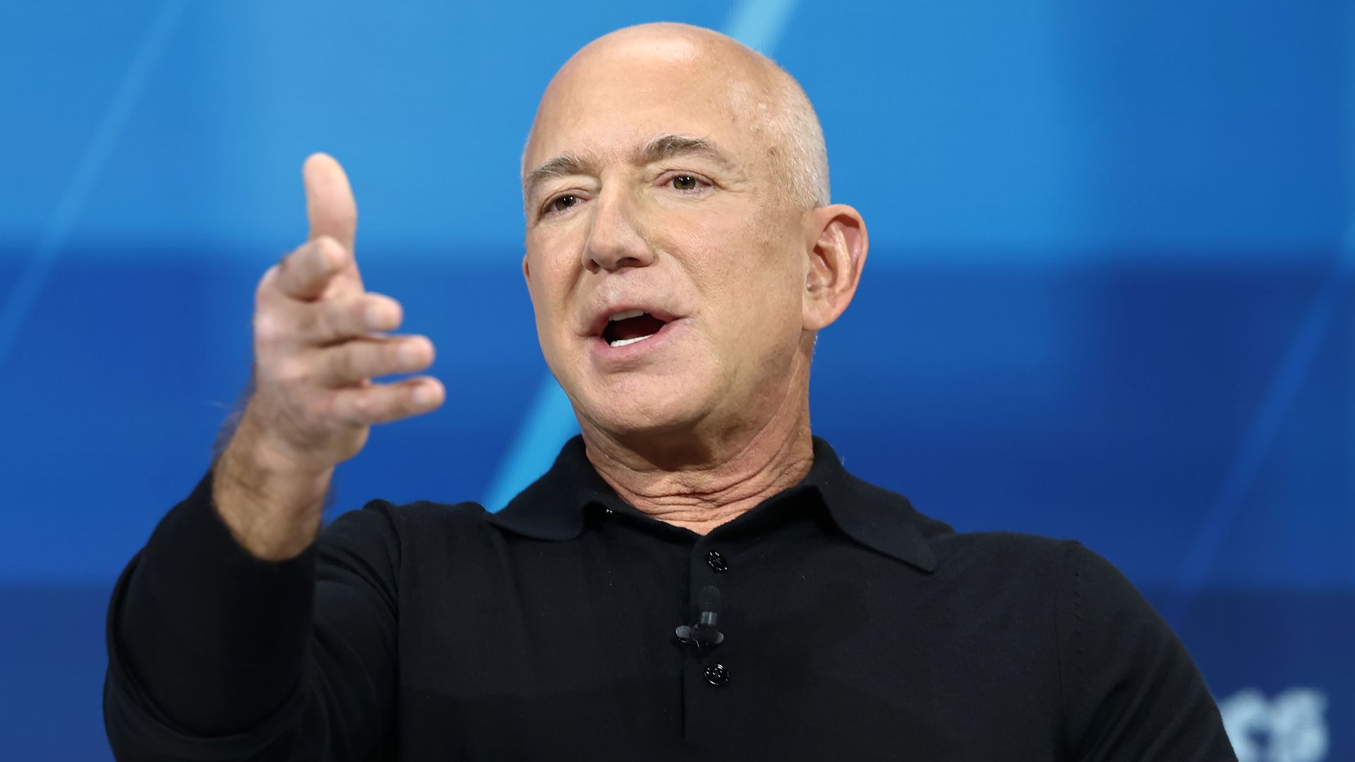 older man, bald, wearing a black shirt talking on stage with a blue background