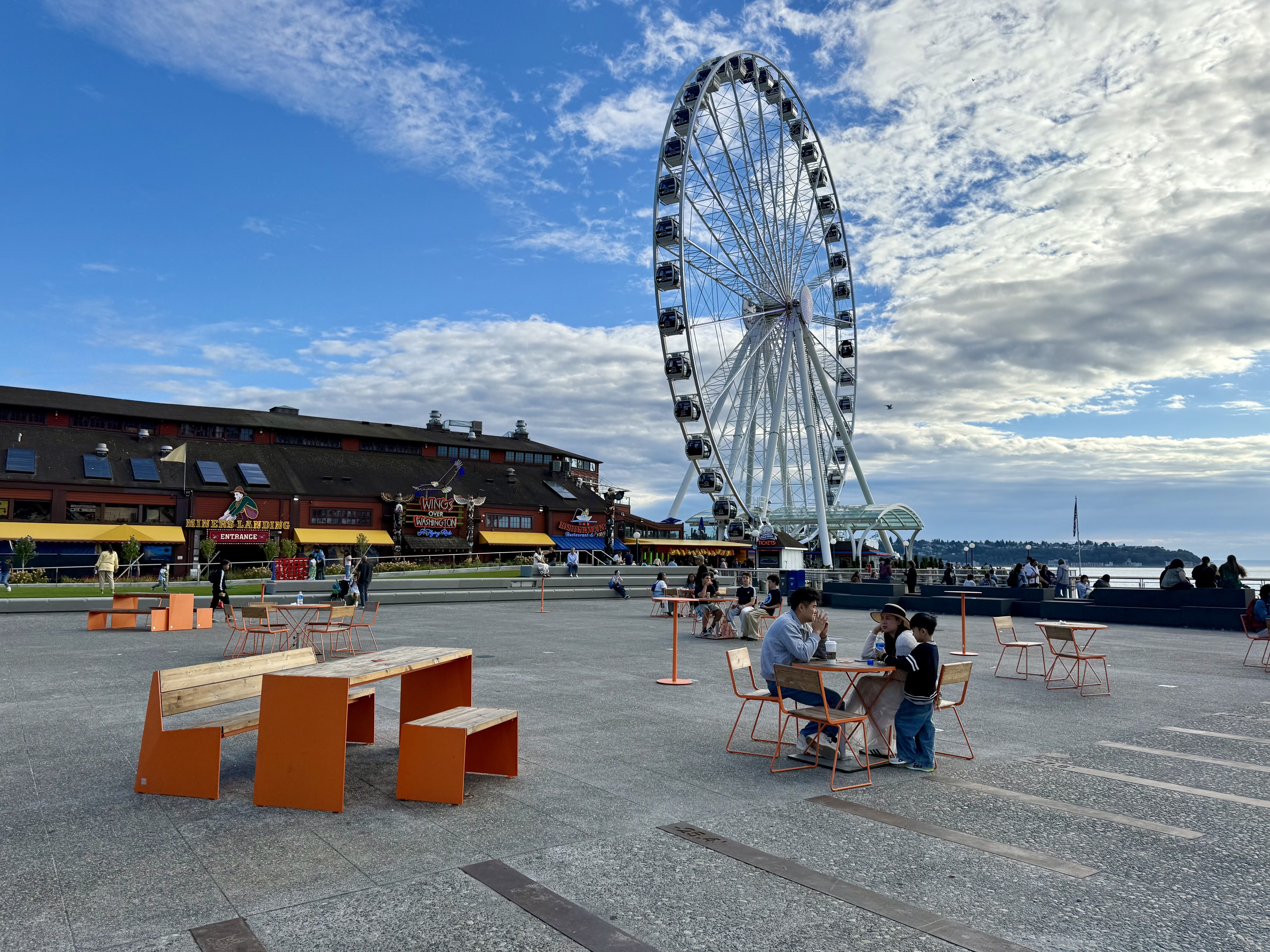 People sitting at orange and wood tables near a large Ferris wheel by the waterfront and a building with neon signs under a partly cloudy blue sky.