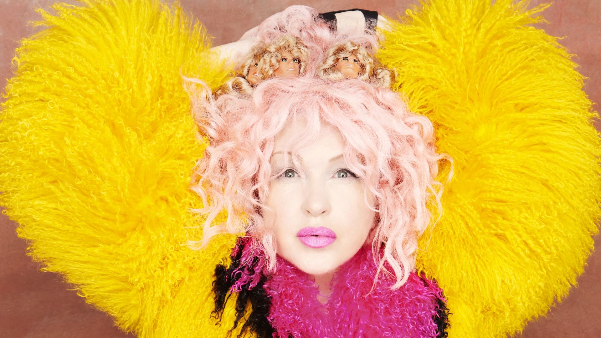 Cyndi Lauper with curly pink hair adorned with two doll heads, wearing a large fluffy coat in yellow, pink, orange, and black, set against a plain brown background.