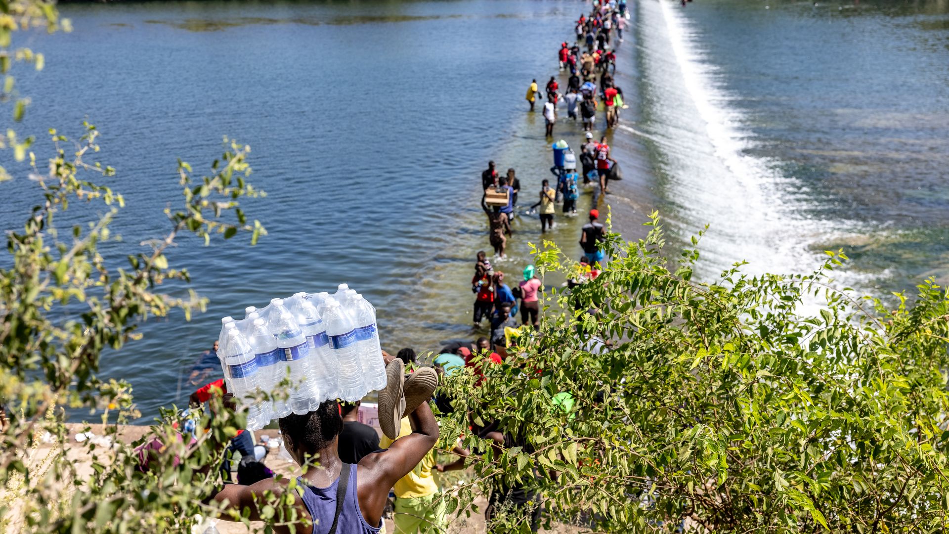 Photo of a stream of people walking across the Rio Grande River carrying belongings and water