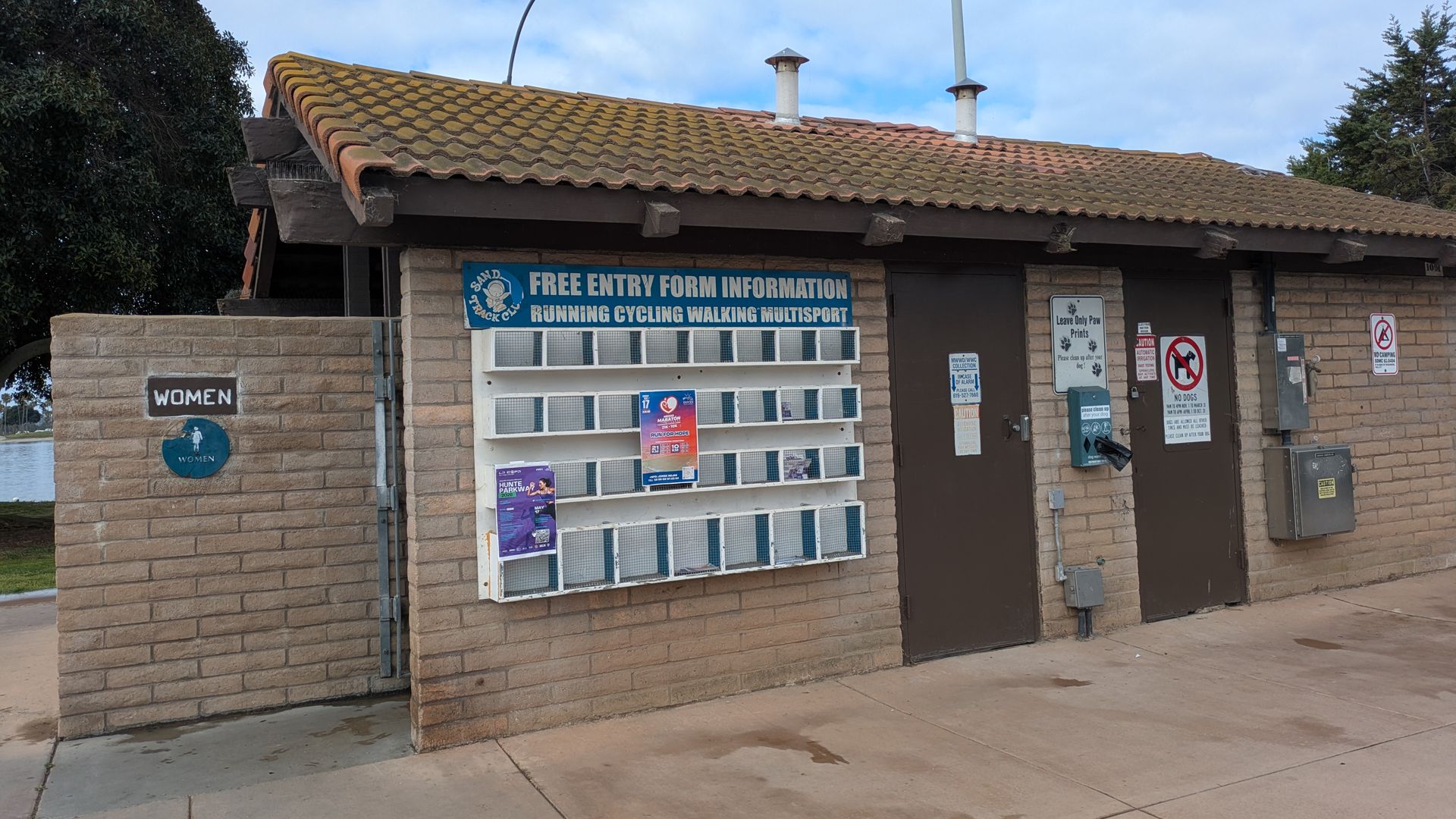 Small brick information booth with a brown tiled roof, two chimneys, and a curved street lamp. A blue poster reads FREE ENTRY above the display; left wall shows a WOMEN sign. Clear sky.