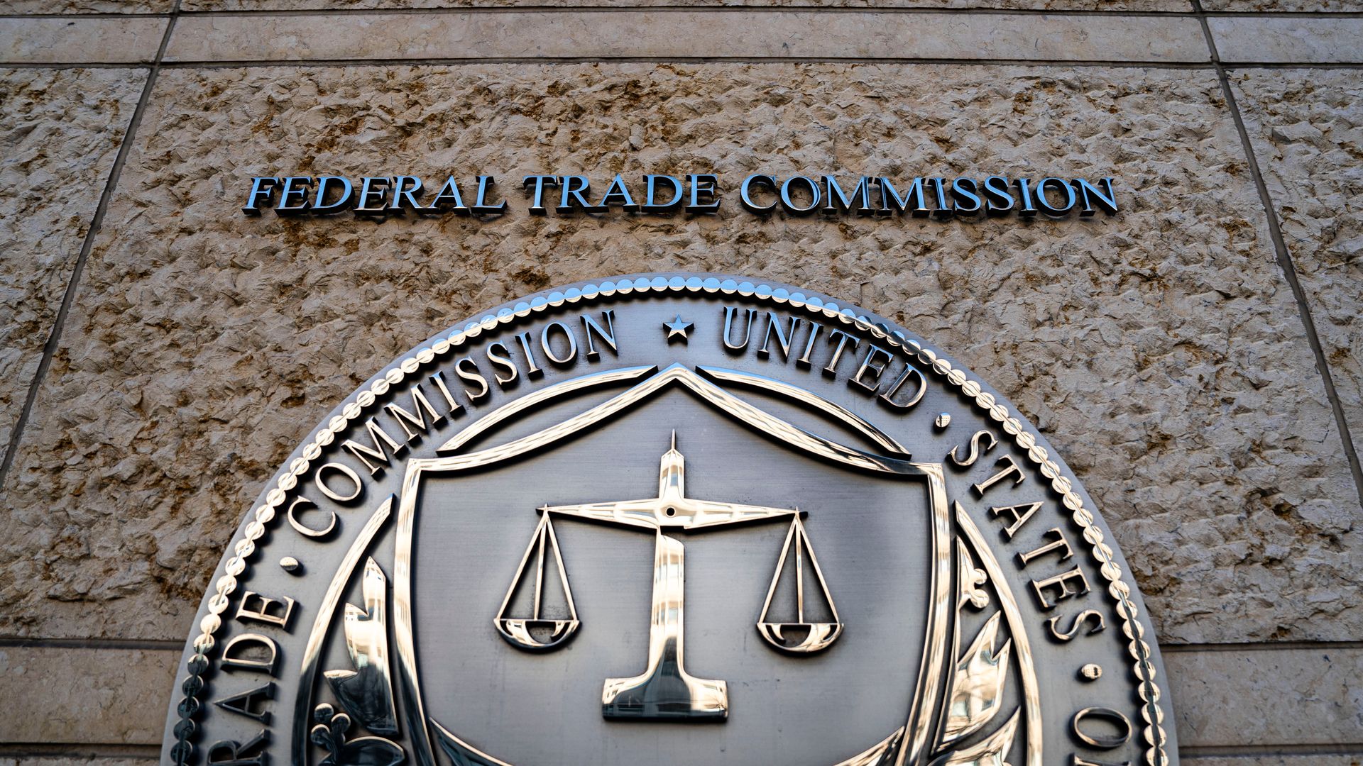 Beige textured stone wall with bold blue letters spelling "FEDERAL TRADE COMMISSION" across. A large circular seal showing the scales of justice is partially visible at the bottom.