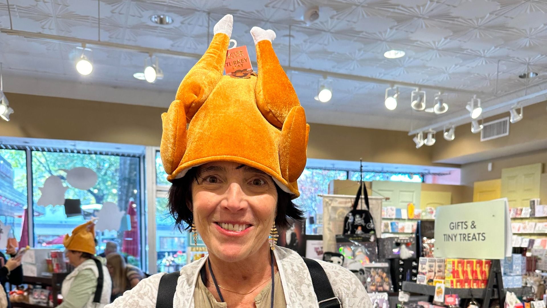 Smiling woman in a store wearing a novelty hat shaped like a roasted turkey, with store shelves and a sign reading "Gifts & Tiny Treats" in the background.