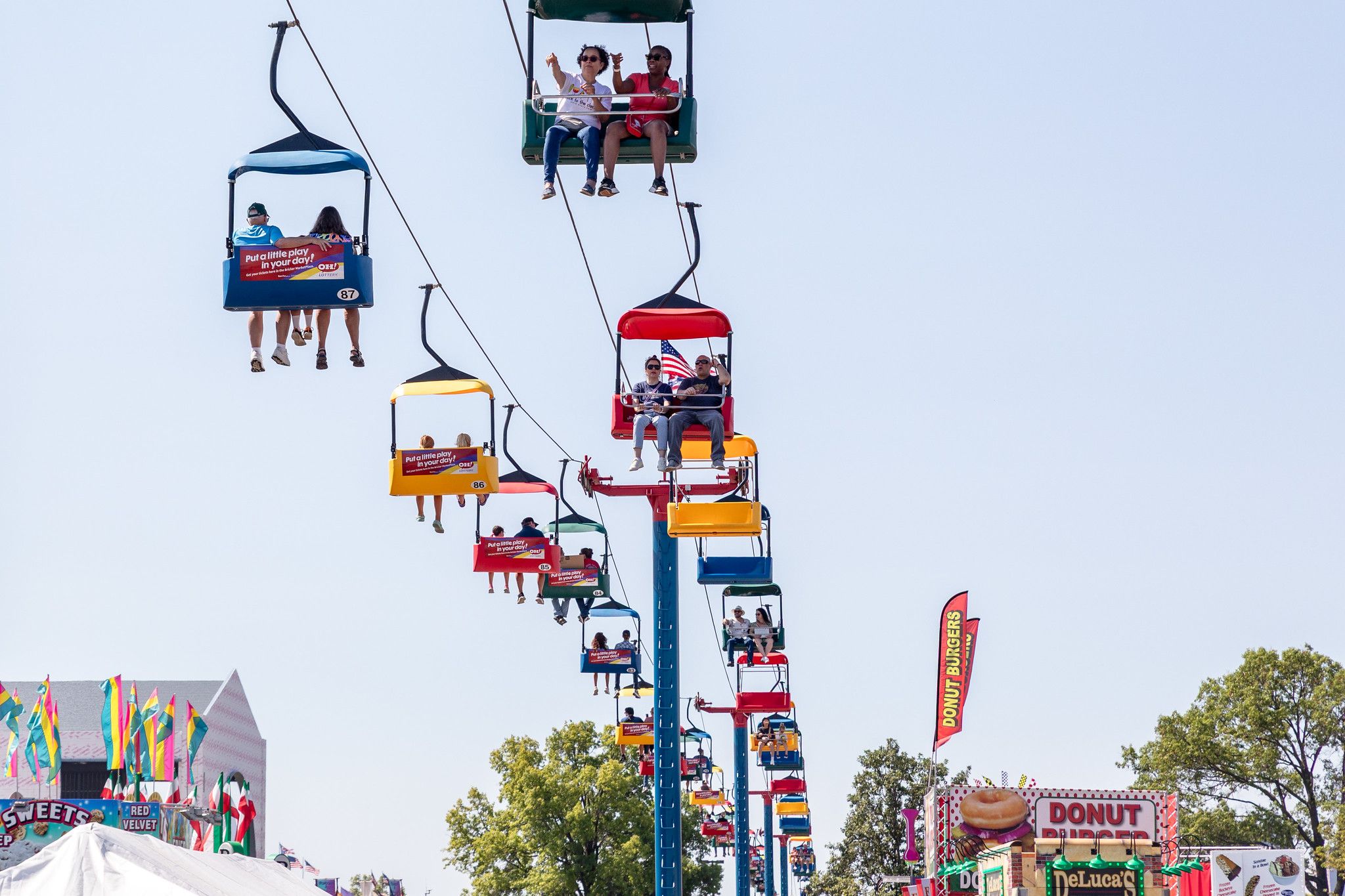 Colorful gondolas rise on a tall carnival ride against a clear blue sky; riders wave and pose. Ground stalls show donuts, bright flags, and striped tents at the fair.