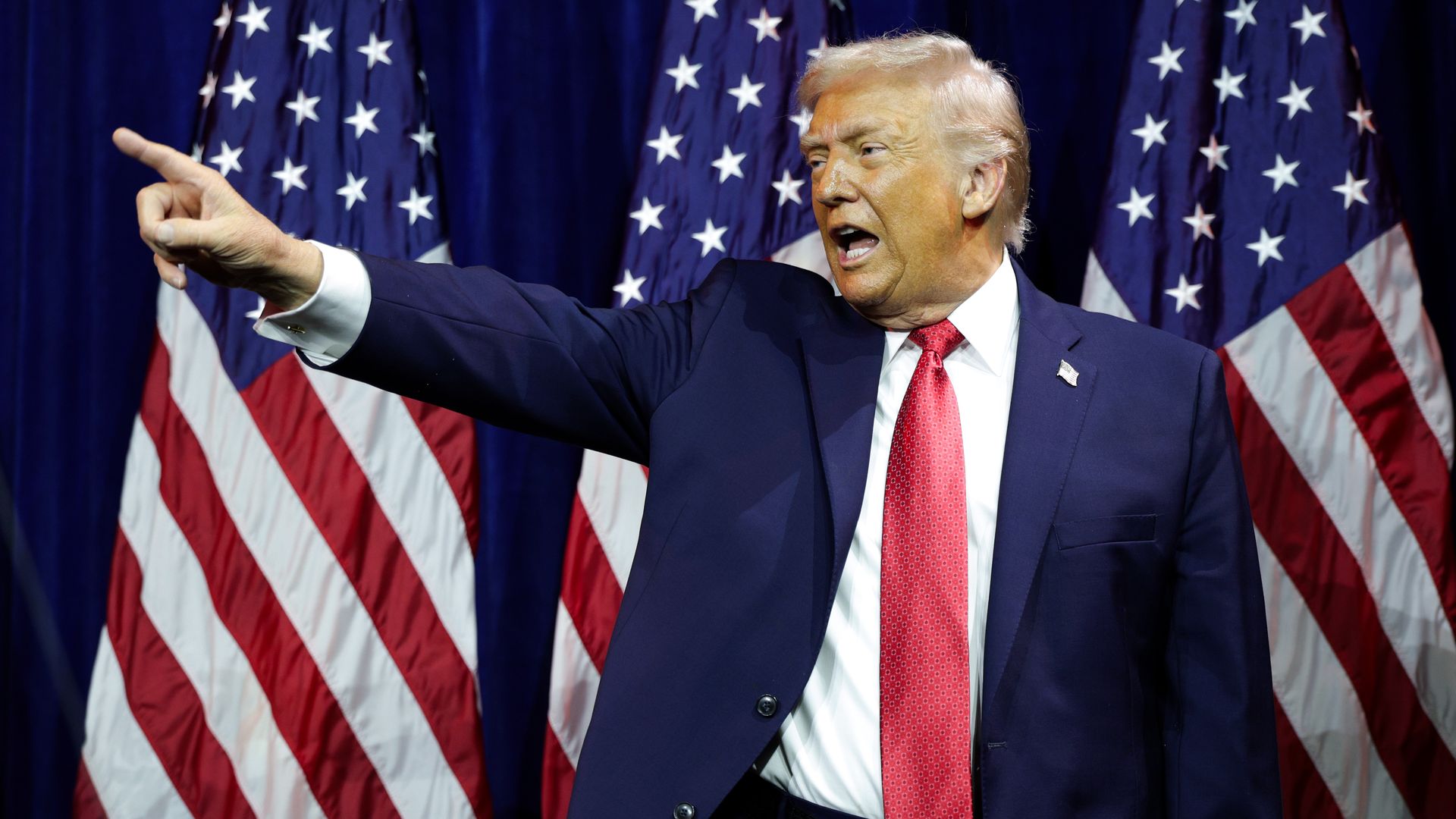 President Trump — wearing a navy suit, a white collared shirt, a red tie and an American flag pin — points while standing in front of three American flags.