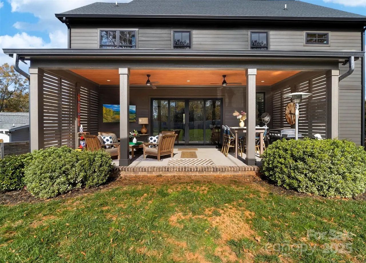 Back view of a gray two-story house with a covered patio featuring wooden furniture with cushions, a dining table, ceiling fans, decorative plants, and green shrubs in the foreground.